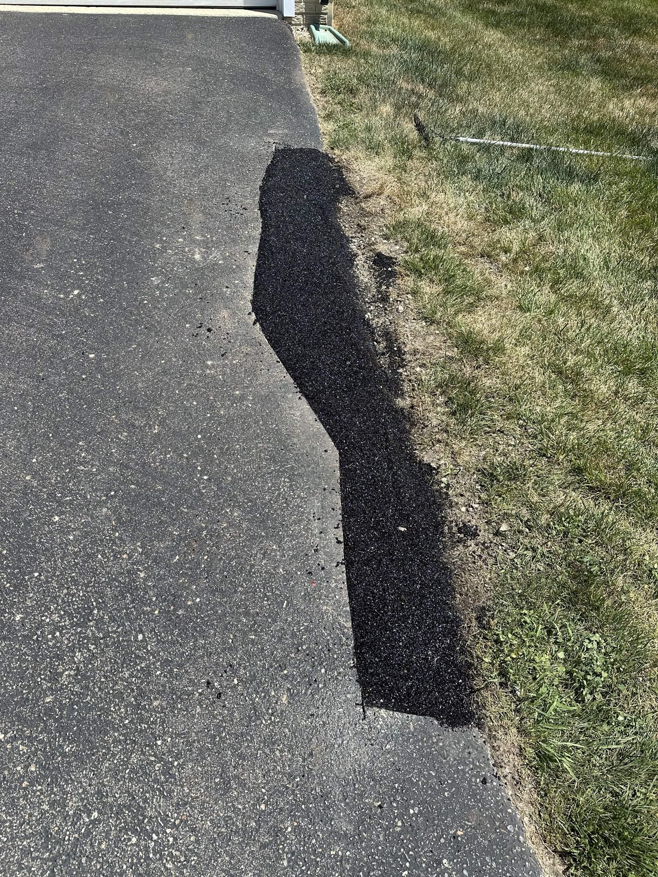 Freshly patched asphalt at the edge of a paved driveway with grass on the nearby lawn.