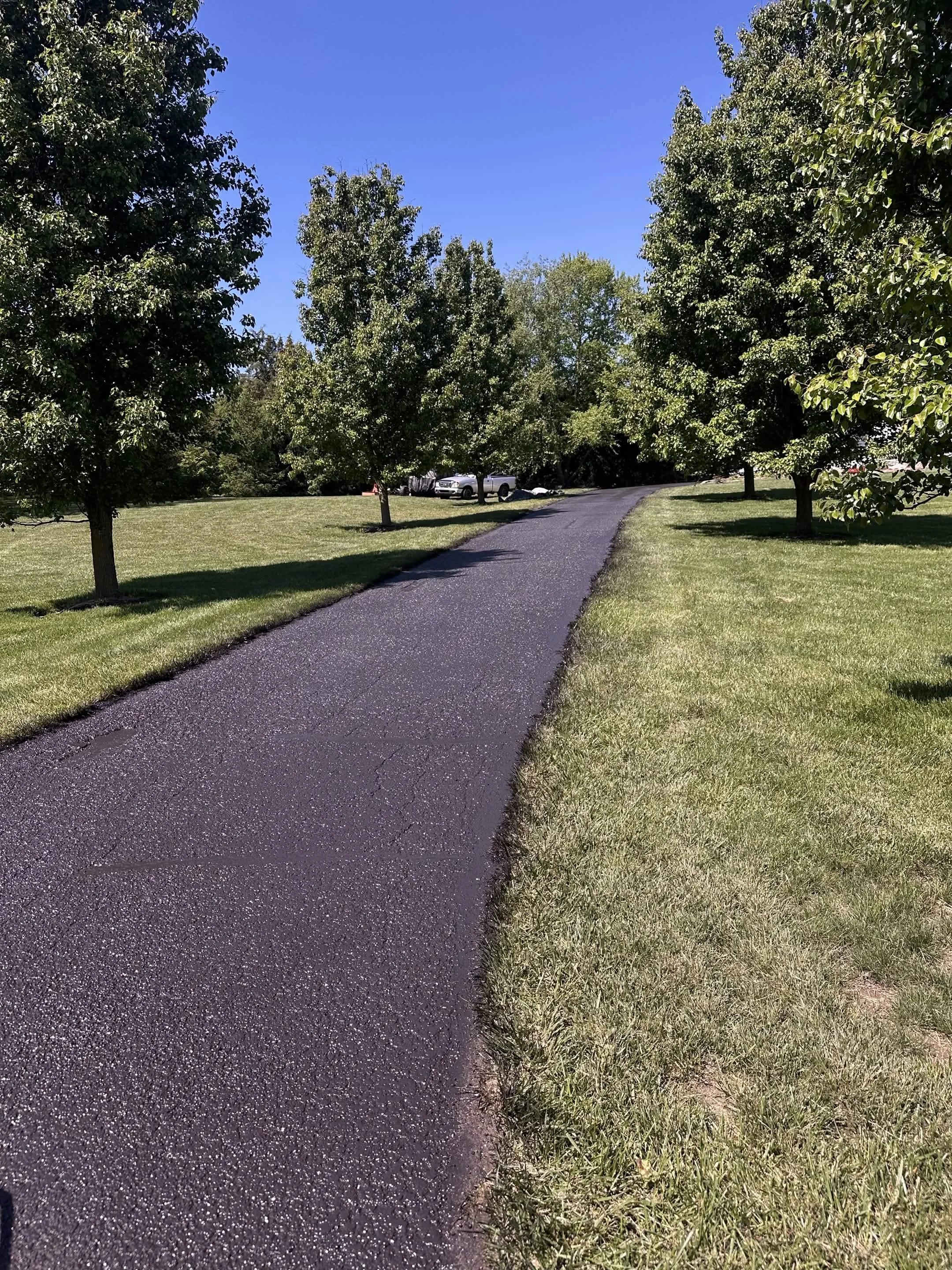 Paved path winding through a grassy lawn with trees on both sides, under a clear blue sky.