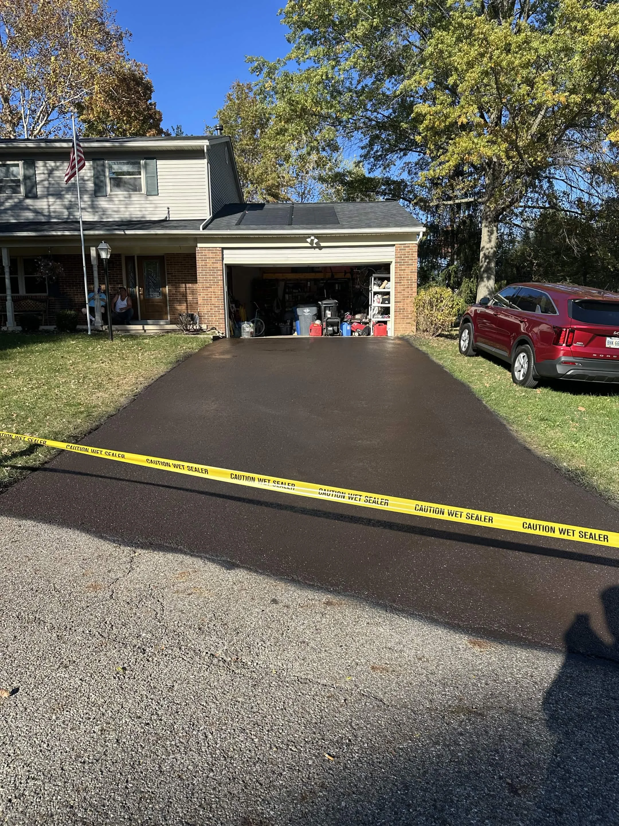 A residential driveway with fresh black asphalt leading to a garage. The driveway is bordered by grass and a concrete sidewalk. A yellow caution tape reading 'Caution Wet Sealer' is across the driveway. In the background, a house with a brick facade,