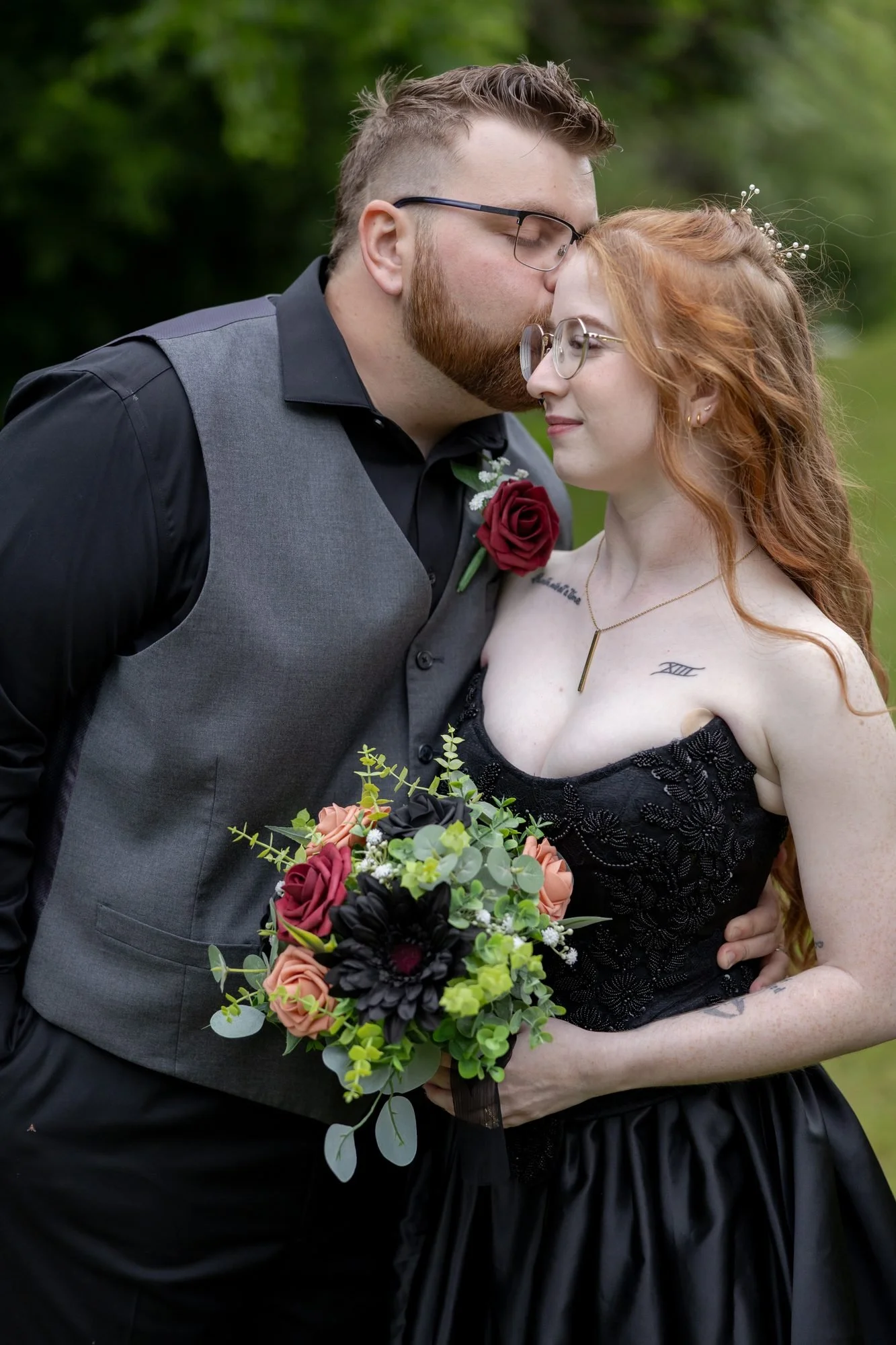 Groom kisses the forehead of a bride in a black wedding dress holding a dark red rose bouquet