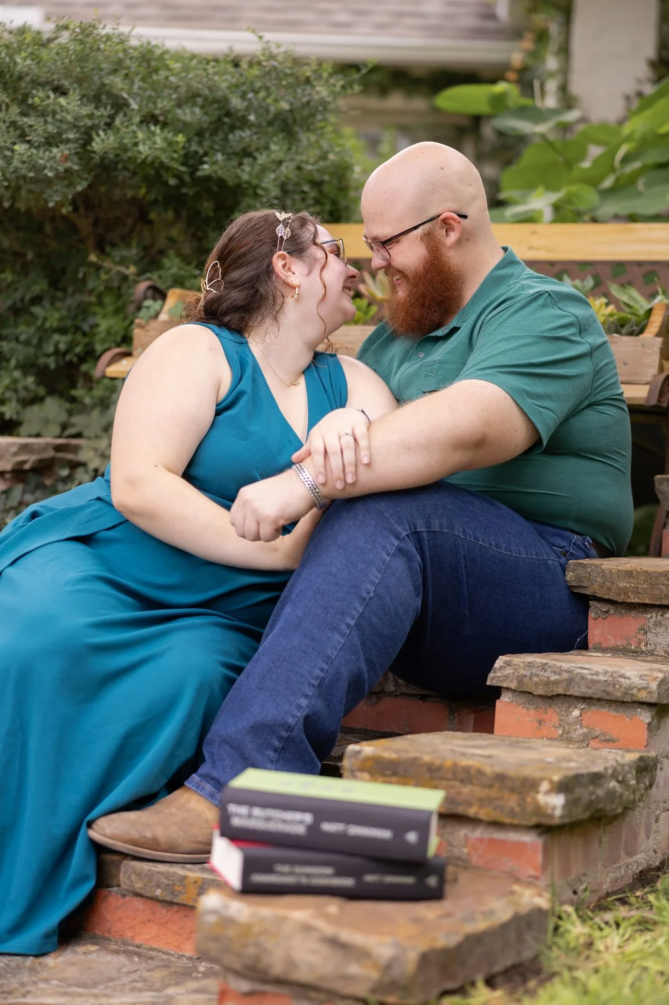 Couple laughing on garden steps with a stack of books during a nerdy engagement session