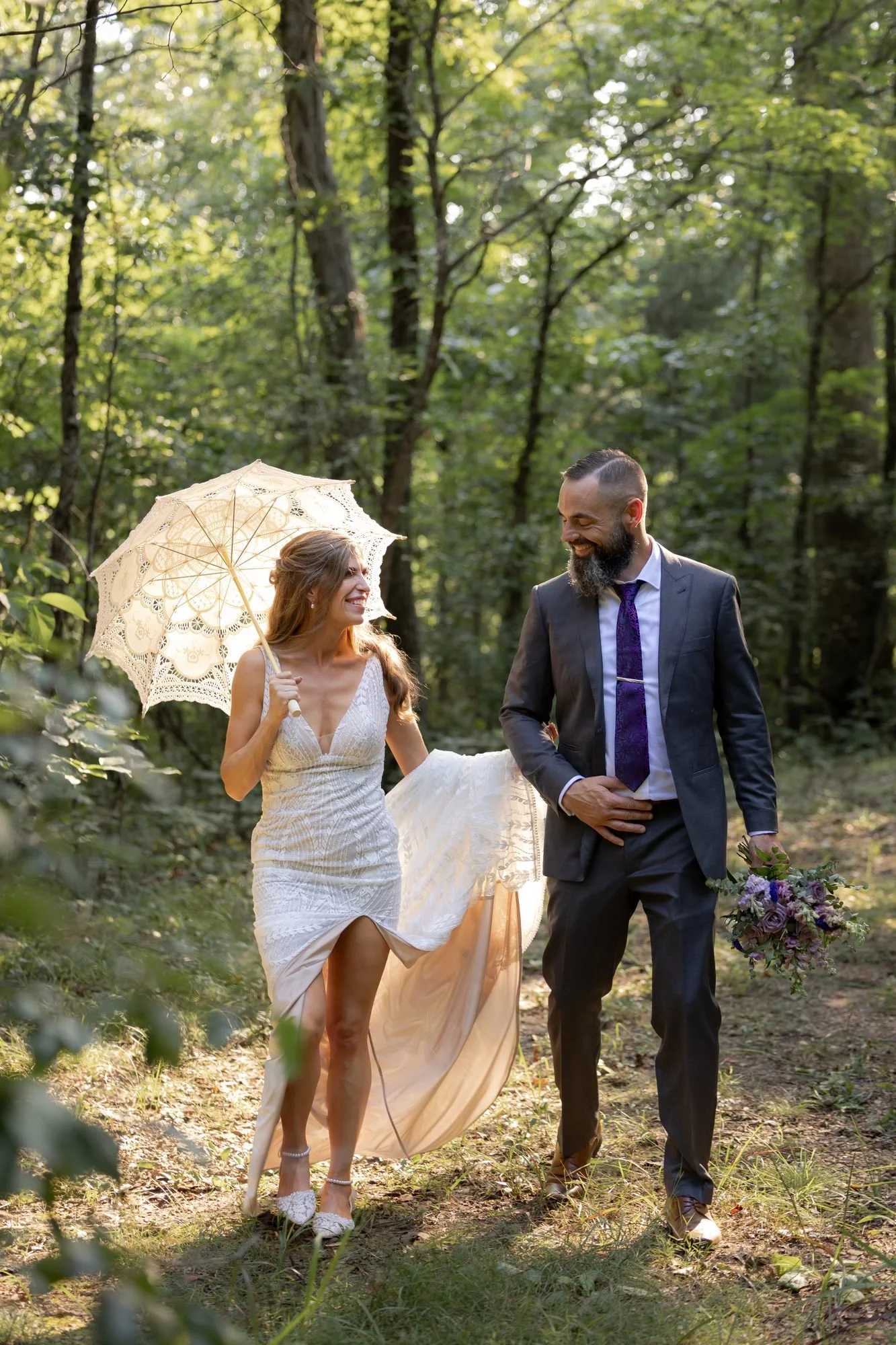 Bride with a lace parasol and groom walking through a sunlit woodland wedding