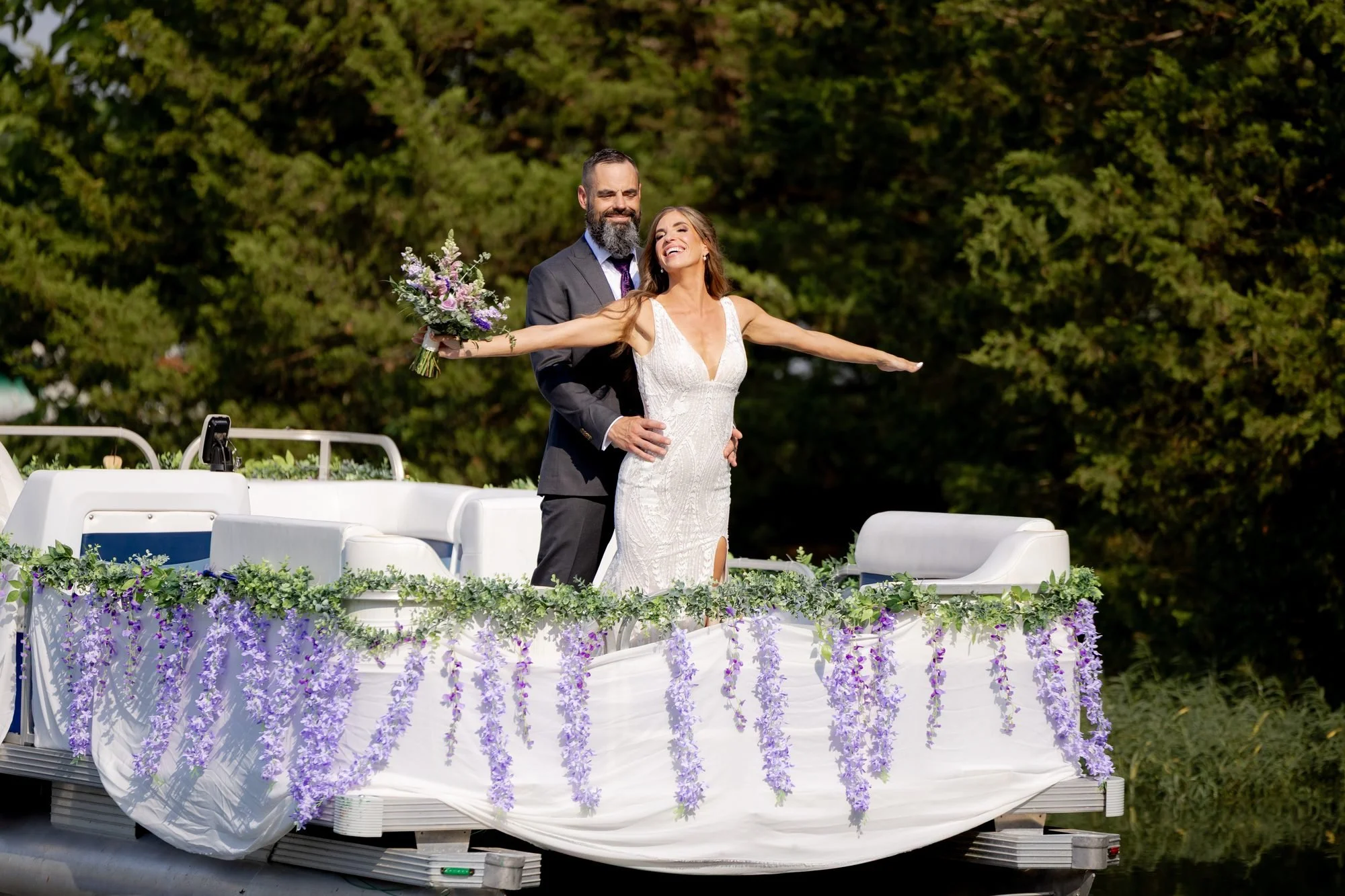 Bride and groom striking a Titanic pose on a flower-covered pontoon boat at their lake wedding