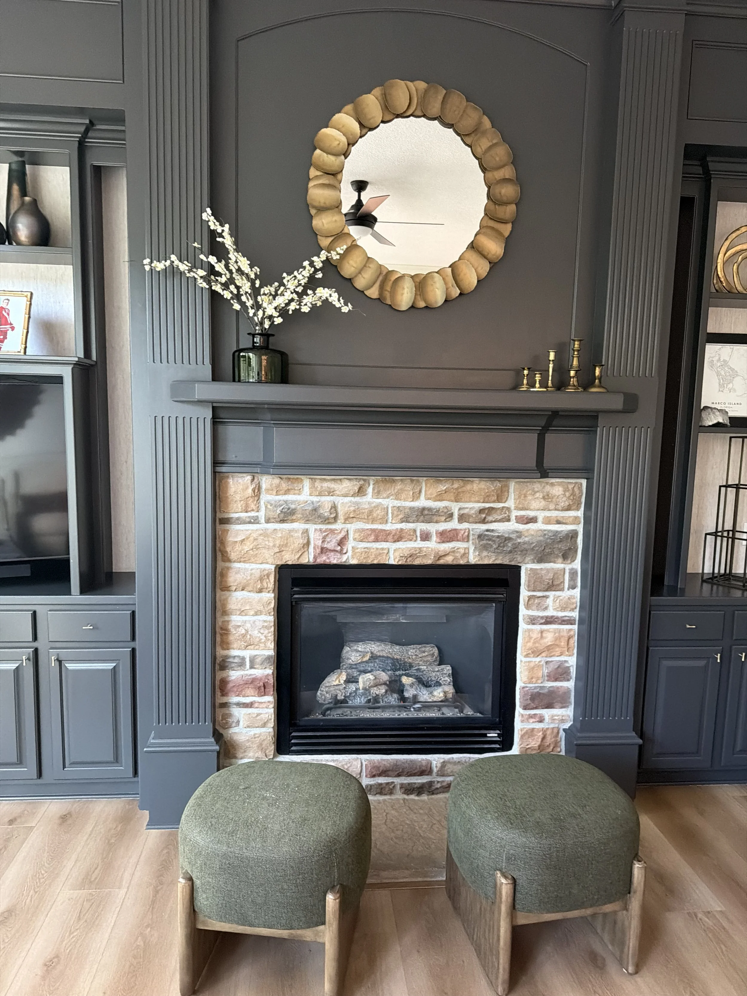 Living room with a fireplace, a decorative mirror with a frame made of bread rolls, a green vase with white flowers on the mantel, and two green upholstered chairs in the foreground.