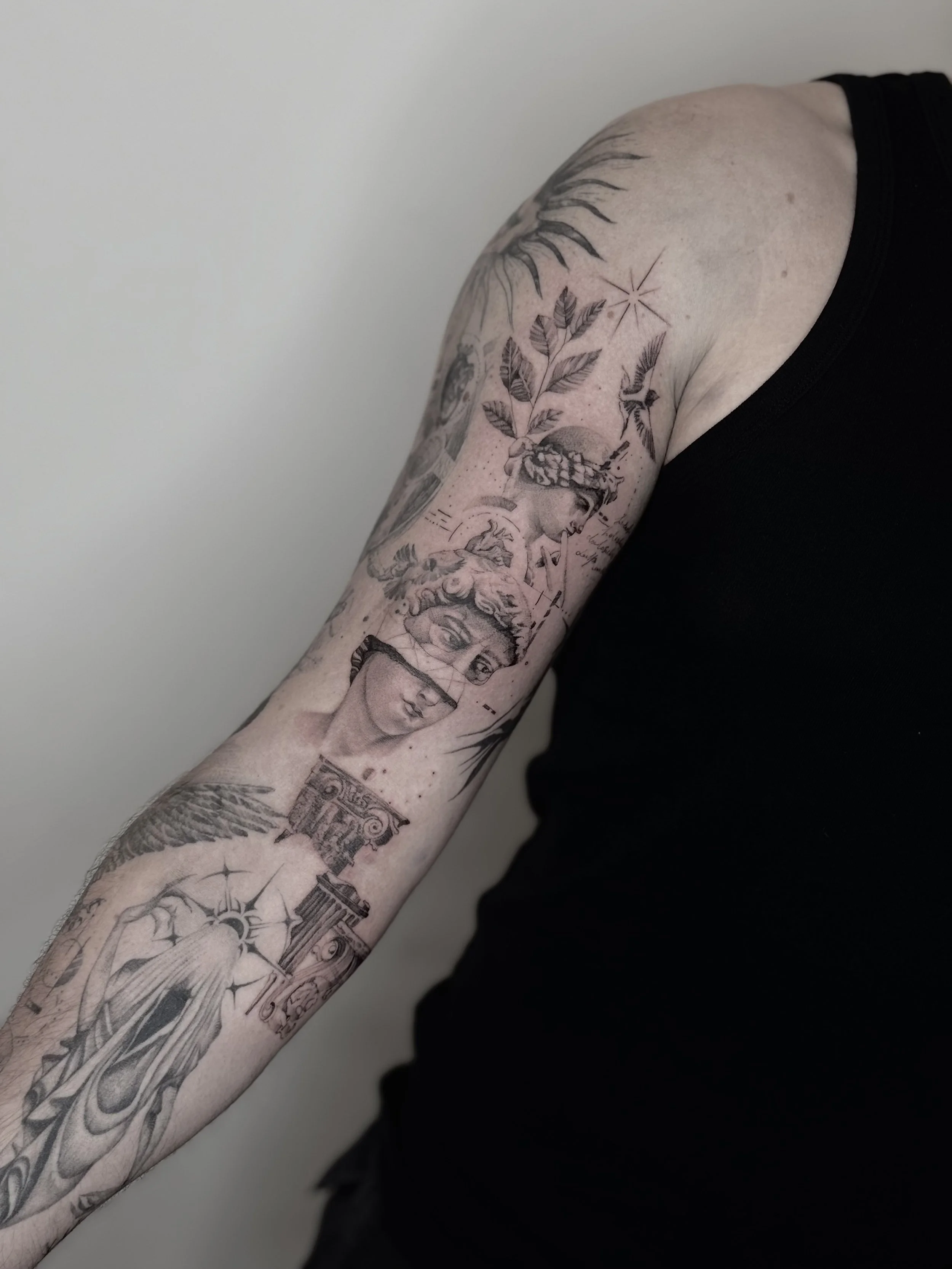 Fine-line tattoo in Paris. A human arm with multiple black tattoos, including a detailed face, various nature-inspired elements, and architectural designs.