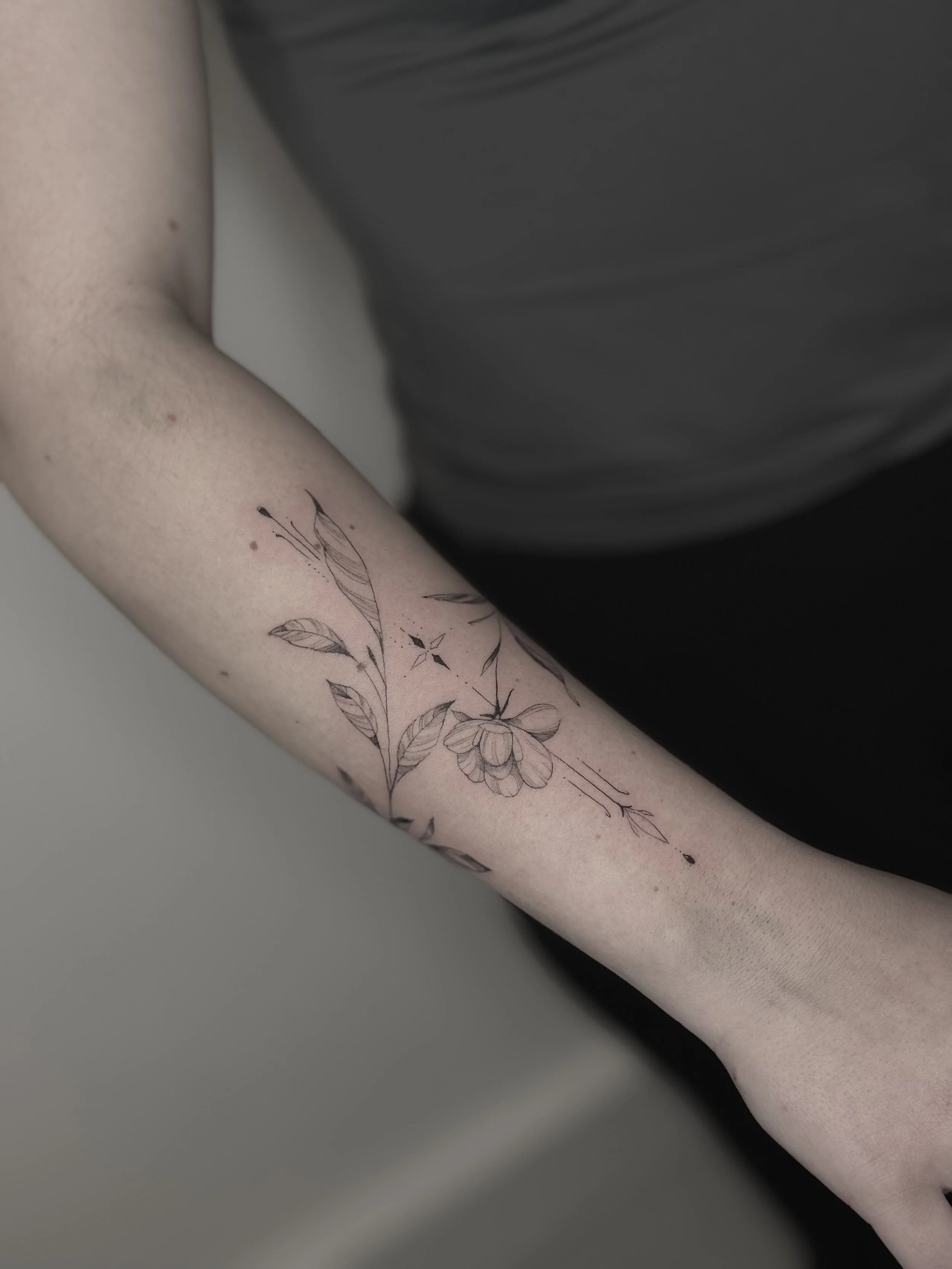A person's arm with a floral tattoo design featuring leaves and flowers in fine-line tattoo style. Paris botanical and fine-line tattoo.