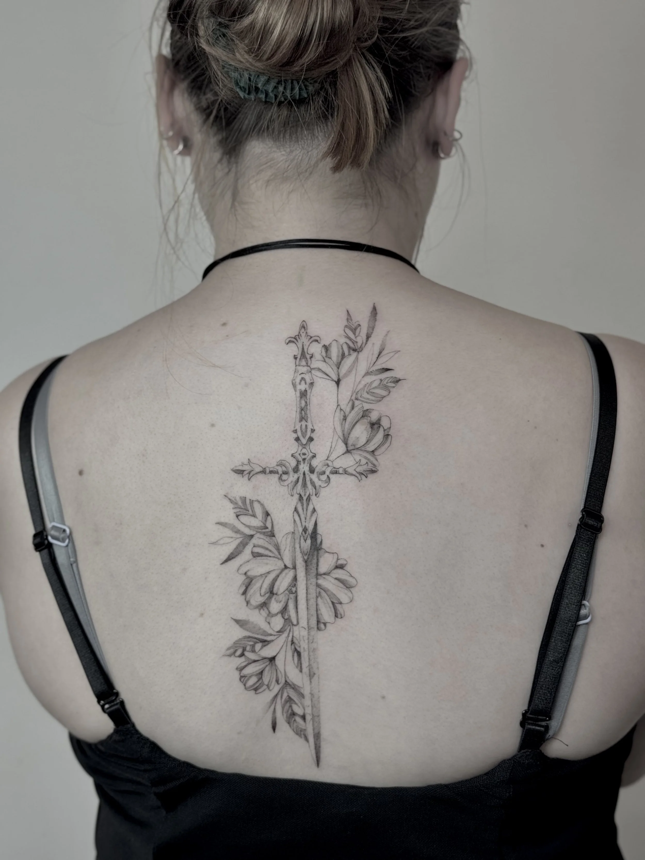 Back of a person's upper body, showing a detailed sword with floral tattoo with leaf elements running down the center of their back.  Discover fine line tattoos at NEXT in Paris. Explore delicate linework, botanical designs and custom compositions.