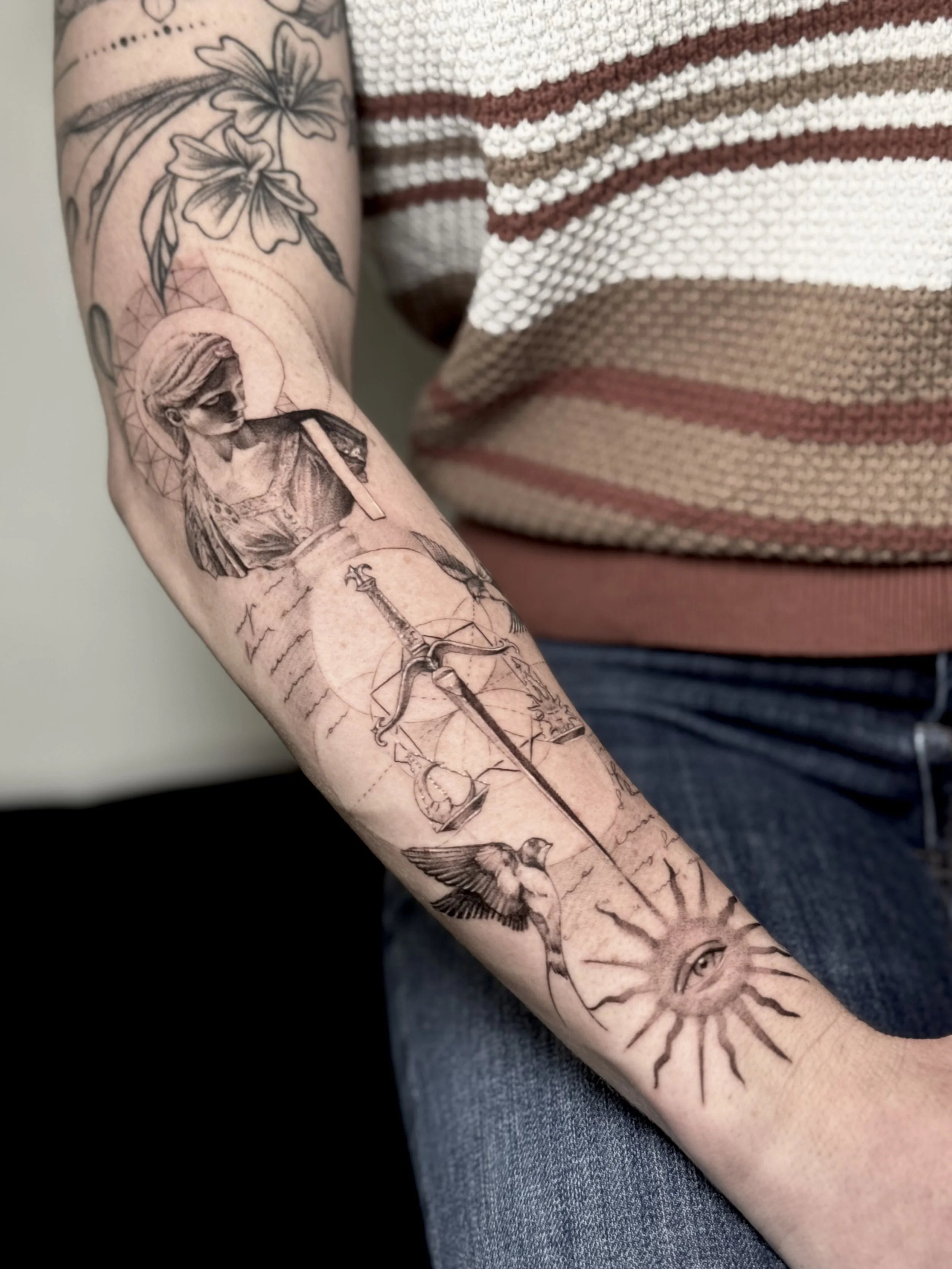 Fine-line tattoo in Paris. A person's forearm tattoo featuring a child with a halo, a sword, flowers, a parrot, an eye, and sun rays.