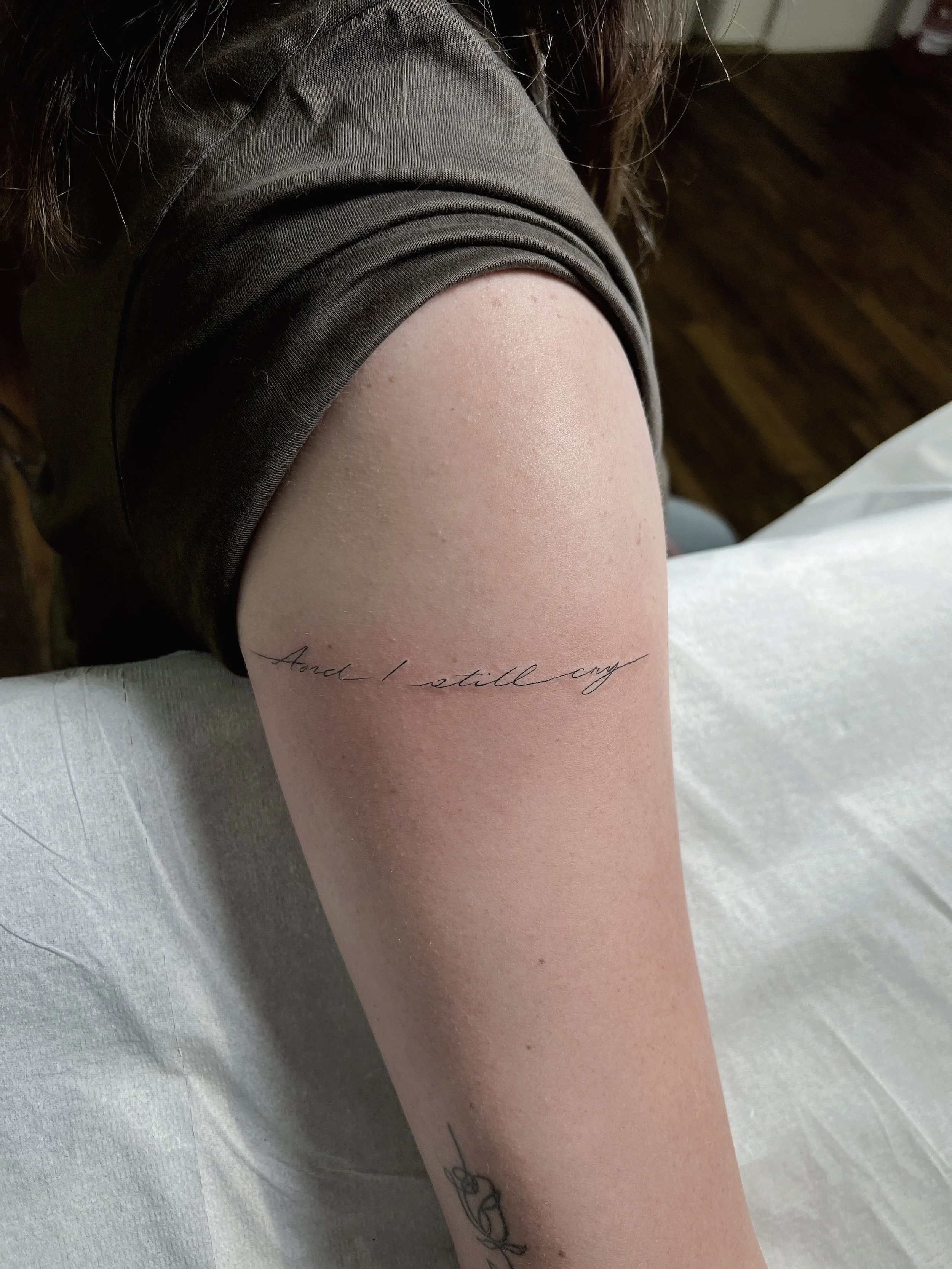 Close-up of a person's upper arm with a tattoo that says 'And I still cry.' The person is wearing a brown or dark-colored short-sleeve shirt, and the background is a wooden floor.