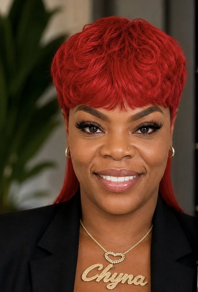 A woman with vibrant red hair styled in a short, curly cut, smiling and wearing a black blazer, gold earrings, and a necklace with a name pendant that says 'Chyna'.