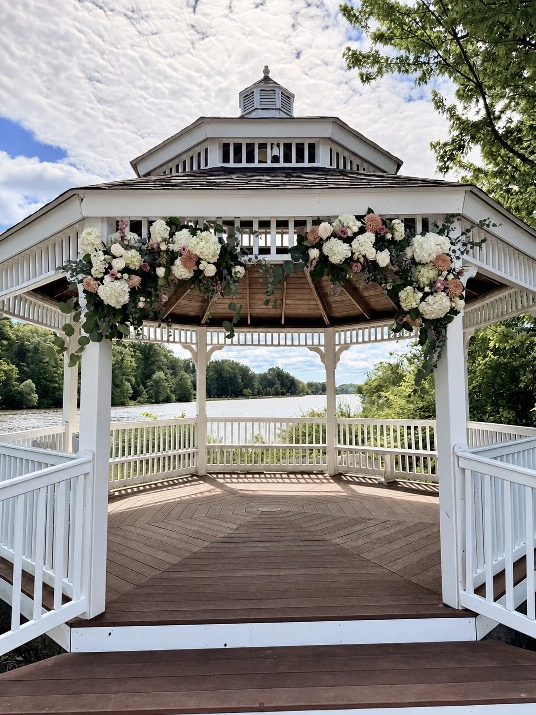 White lakeside gazebo decorated with floral arrangements and overlooking a wooded lake