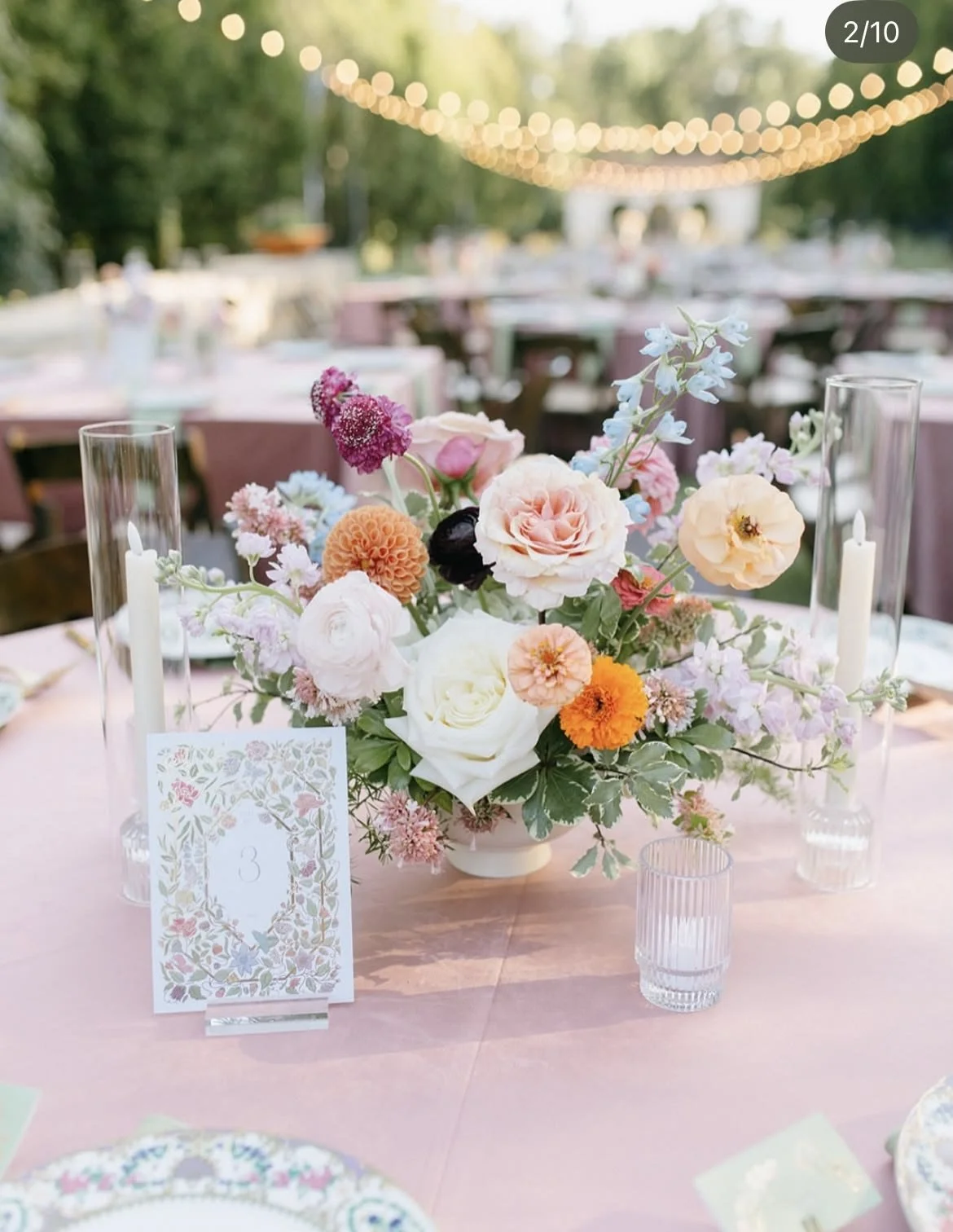 Pastel floral wedding table centerpiece with candles, table number three, and string lights outdoors