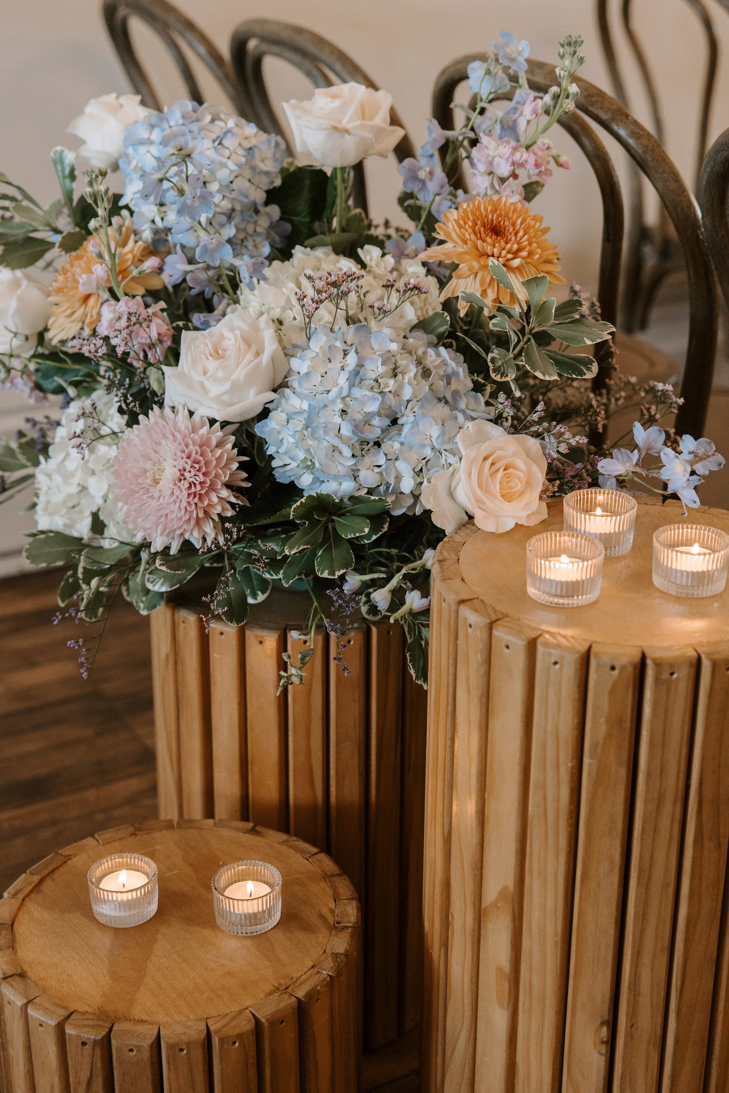 Colorful floral arrangement with roses, hydrangeas, chrysanthemums, and greenery displayed on rustic wooden tables beside lit candles.