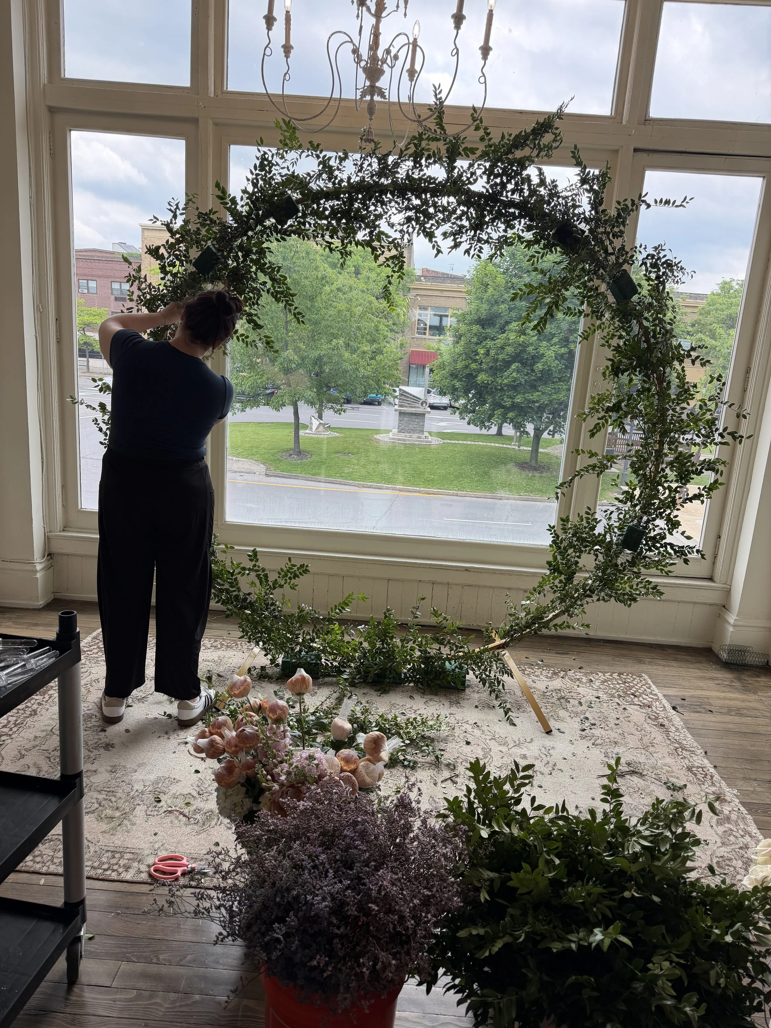Florist arranging a large greenery arch beneath an ornate chandelier in front of tall windows