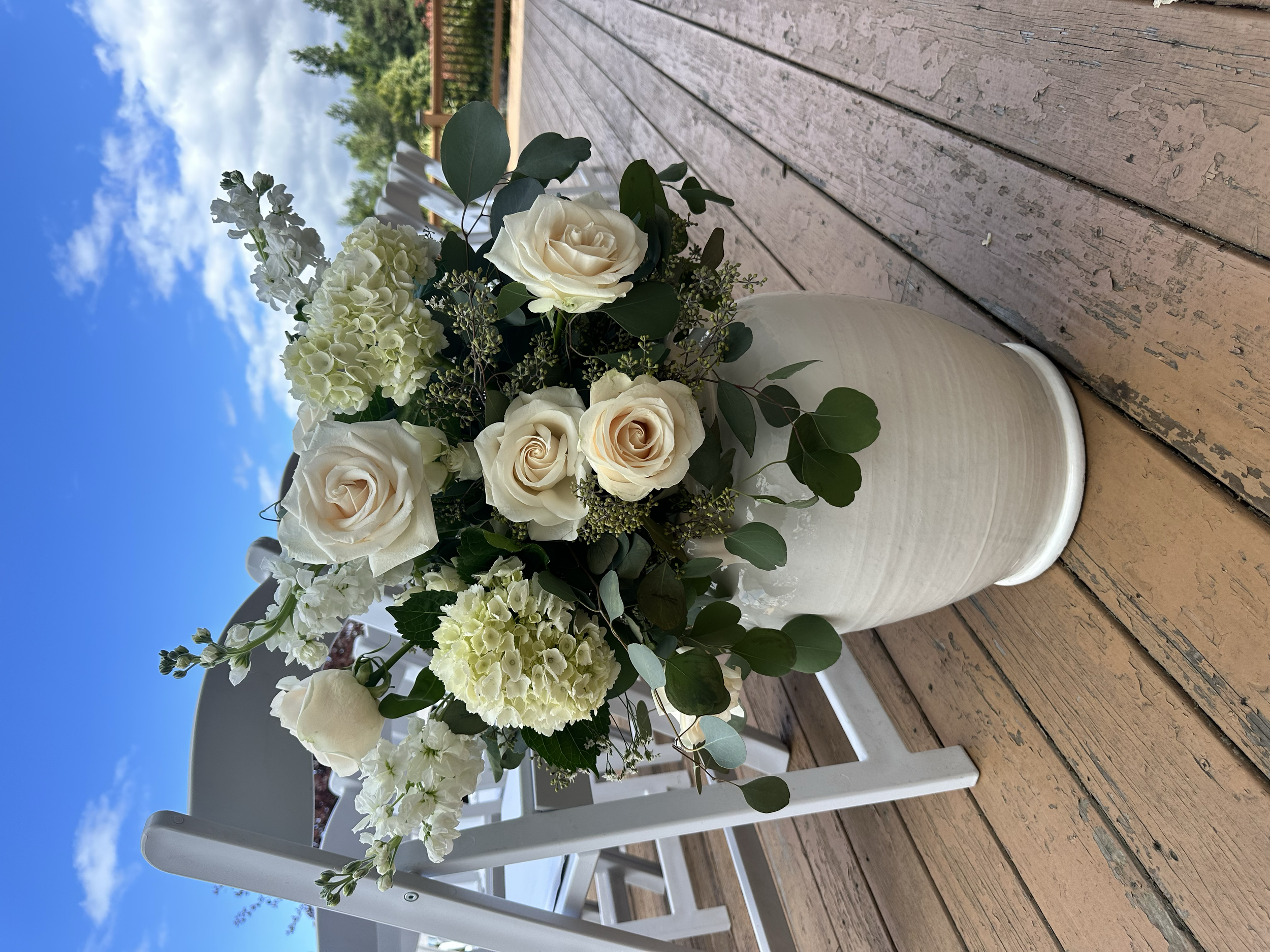 White rose and hydrangea floral arrangement with greenery in a white vase on a wooden deck