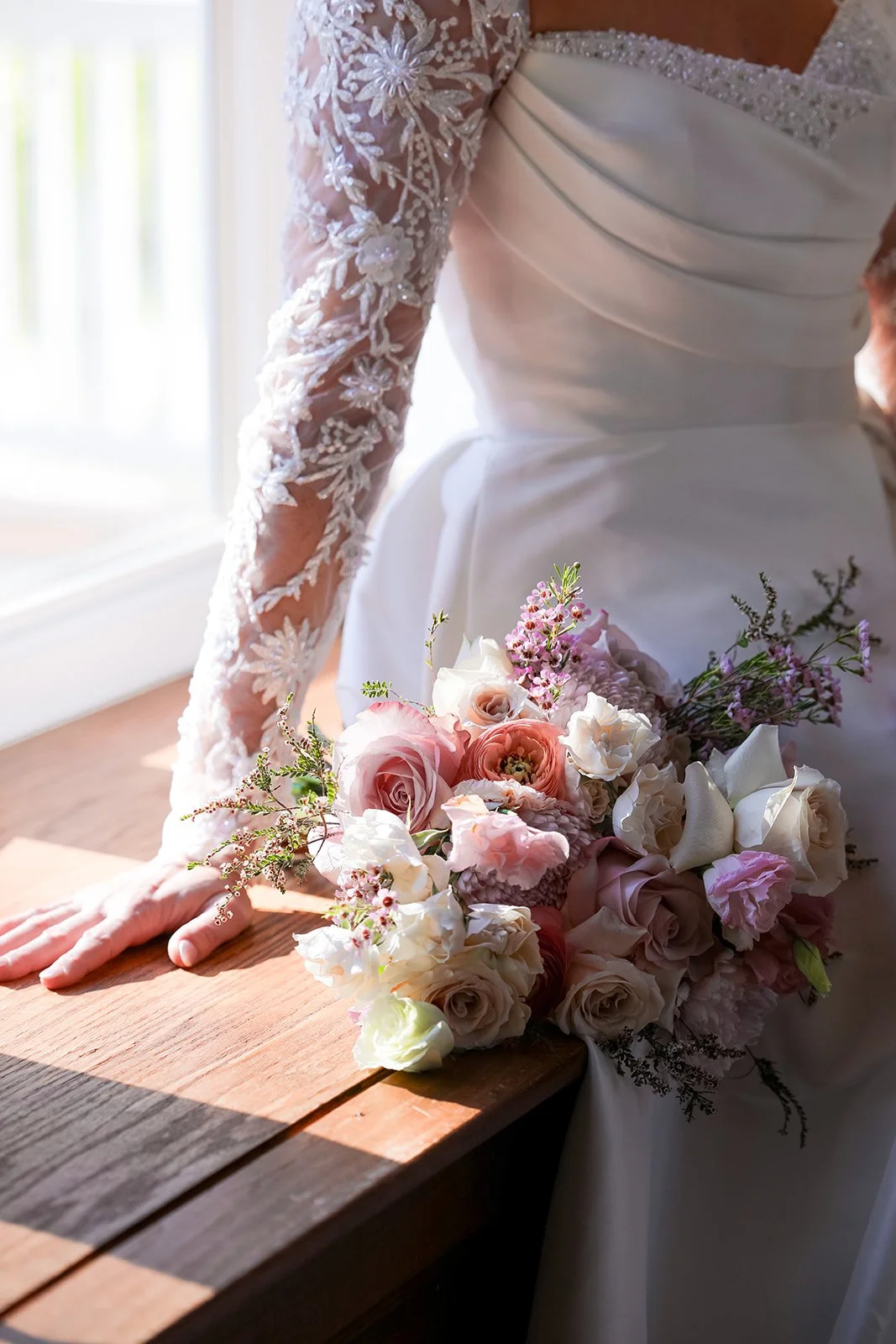 Bride in a white lace-sleeved wedding dress holding a pastel flower bouquet beside a sunlit wooden table