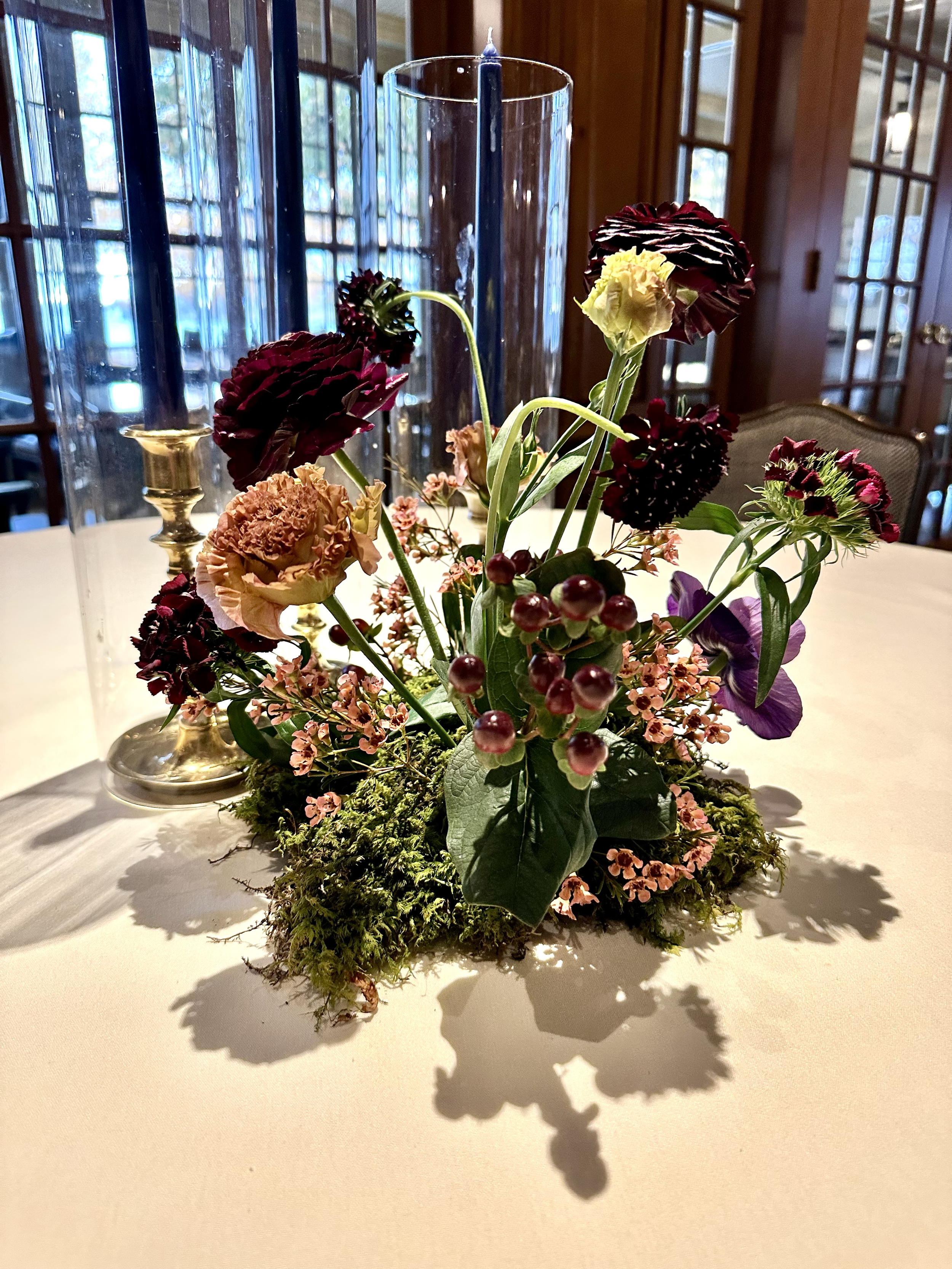 Colorful floral arrangement with burgundy, yellow, peach, and purple flowers, greenery, berries, and moss on a dining table beside candleholders.