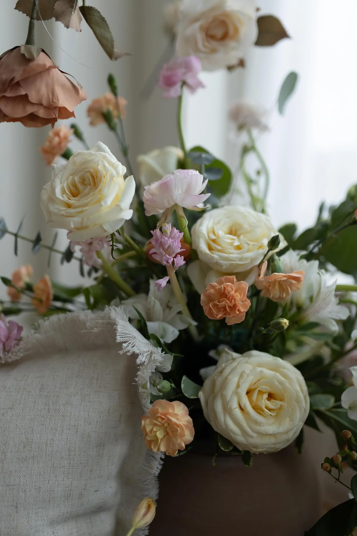 Vintage-style floral arrangement with cream roses, peach carnations, pale pink flowers, and greenery in soft natural light