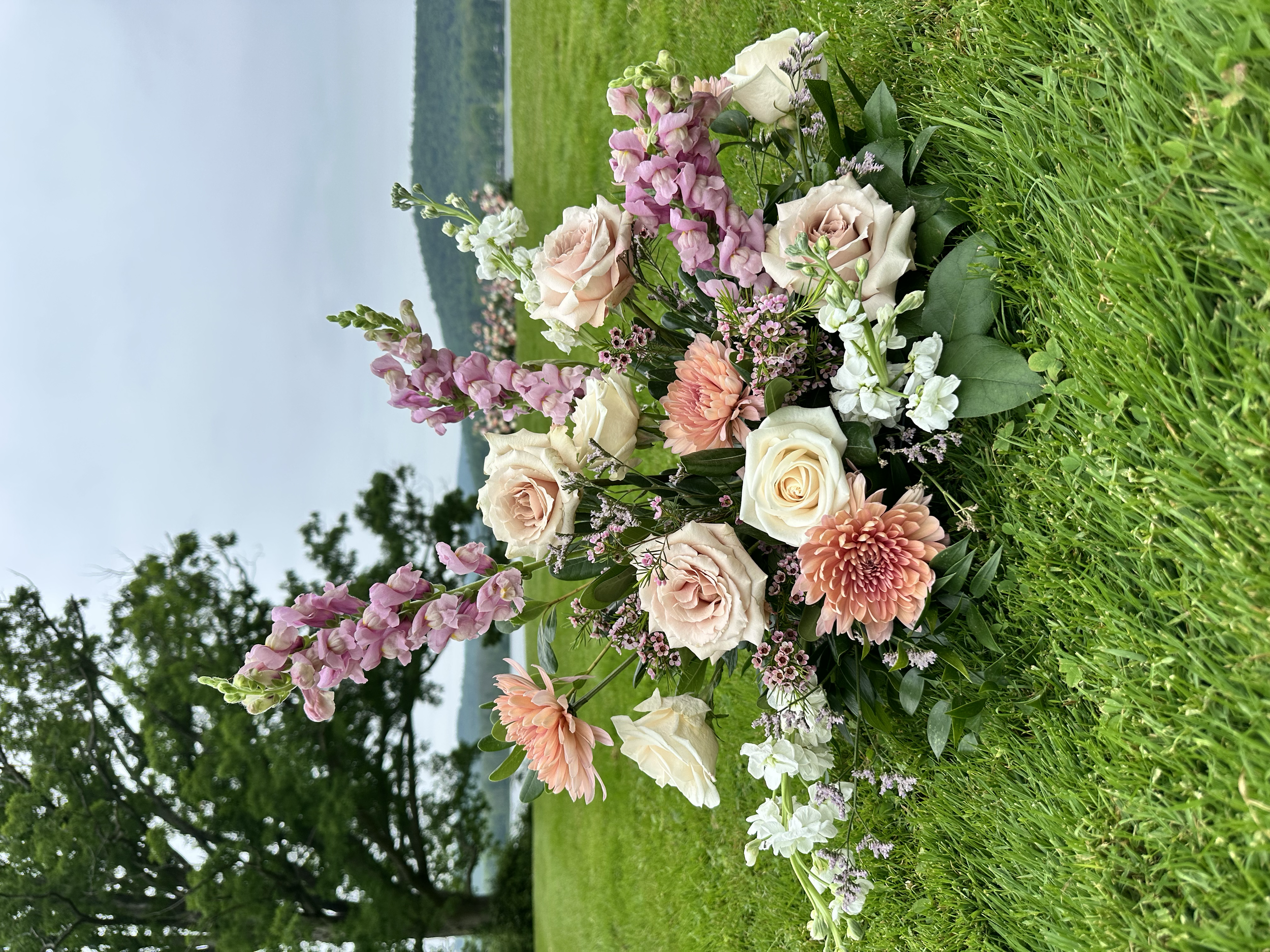 Pastel pink and cream flower arrangement with roses, chrysanthemums, and snapdragons on a lakeside lawn
