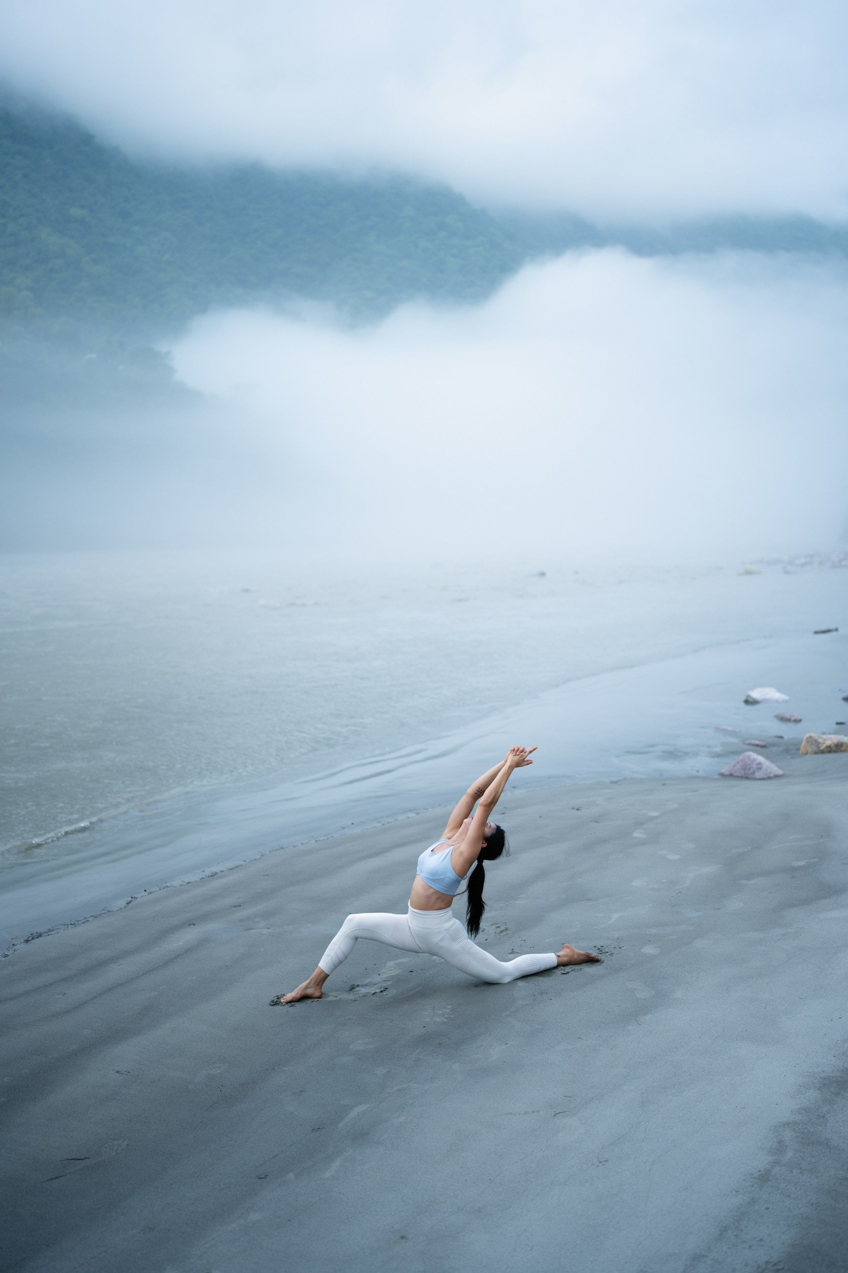 A woman practicing yoga on a foggy beach, performing a low lunge pose with arms raised upward, wearing white workout clothing.