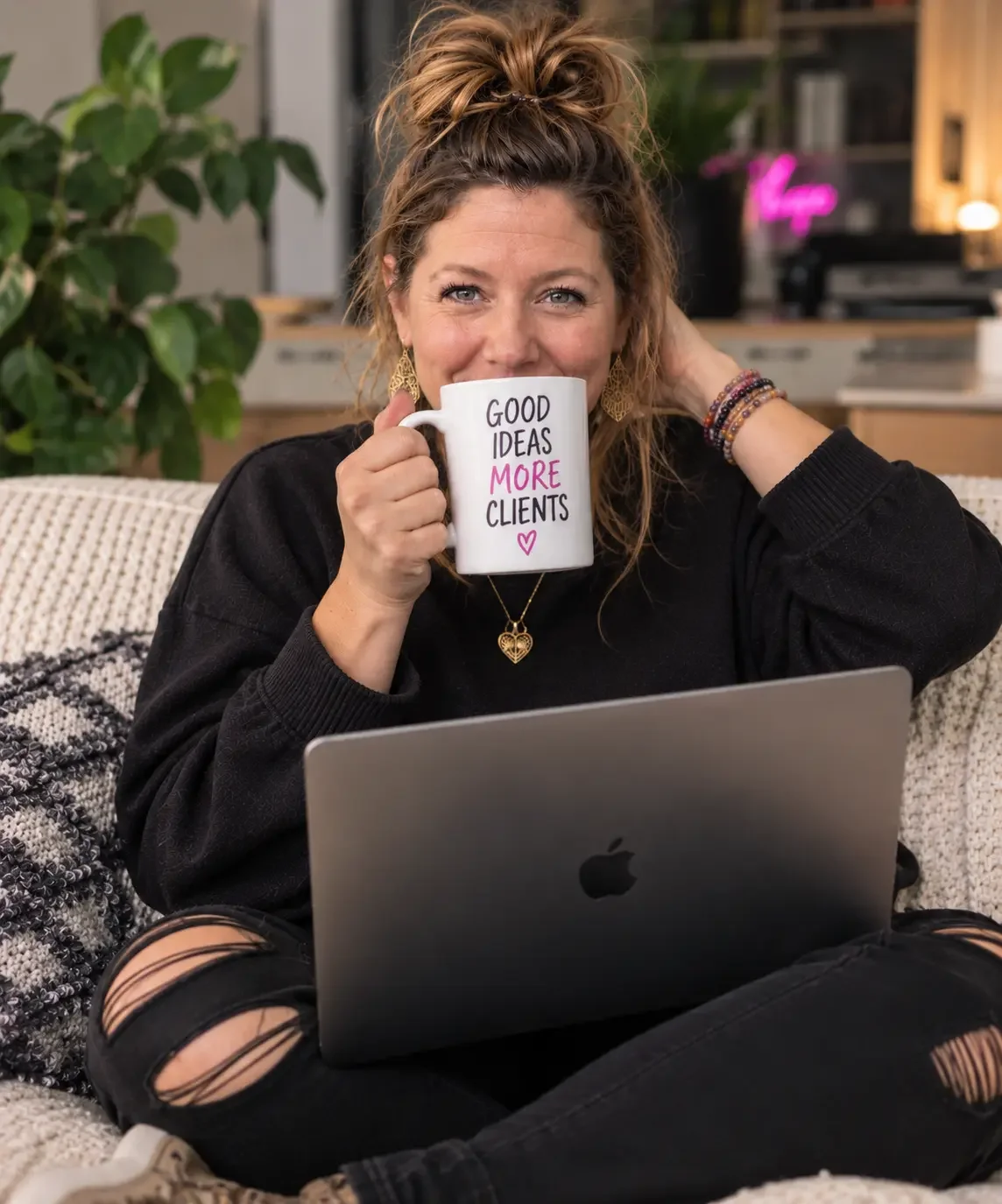 A woman sitting on a cozy couch, drinking from a mug that reads 'Good ideas more clients,' with a laptop on her lap, wearing ripped black jeans and a dark sweater, inside a stylish living room with a plant and background decor.
