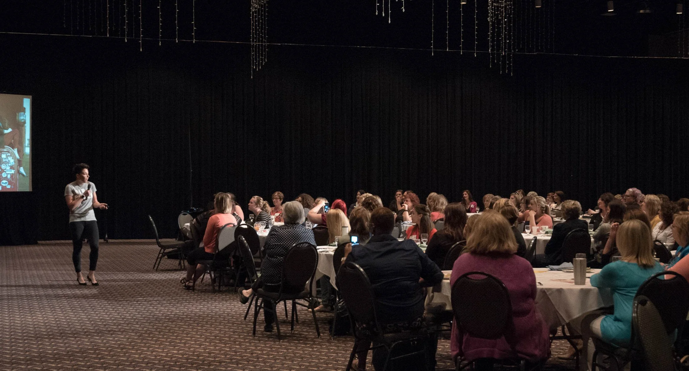 Mel presenting to a packed conference center with the  audience seated at round tables. Mel holds a microphone and stands near a projection screen displaying a slide. The audience consists mainly of women