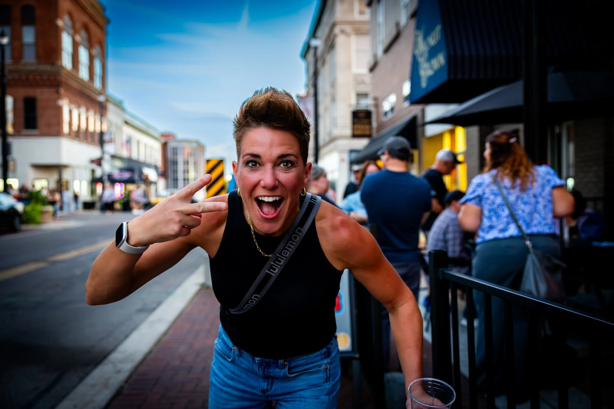 A woman with short brown hair making a peace sign with her hand and smiling broadly in an outdoor urban setting, surrounded by people sitting at tables and buildings on a busy street.