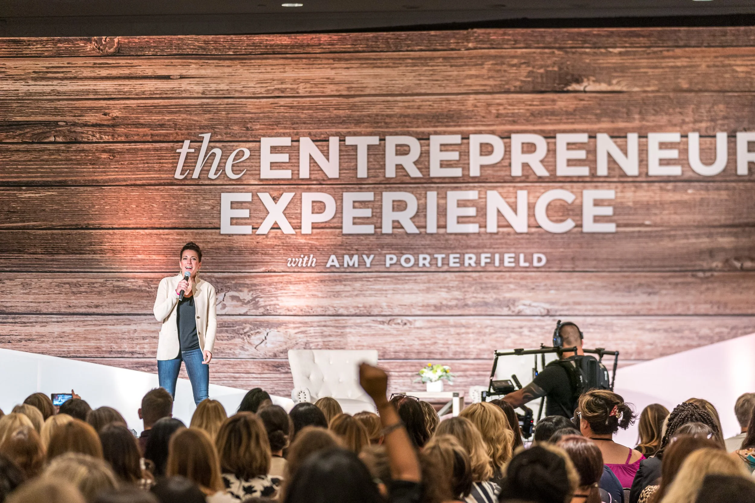 A woman speaking into a microphone on stage at an event called 'The Entrepreneur Experience' with an audience watching. The backdrop features a large wooden design with the event's name and host, Amy Porterfield.