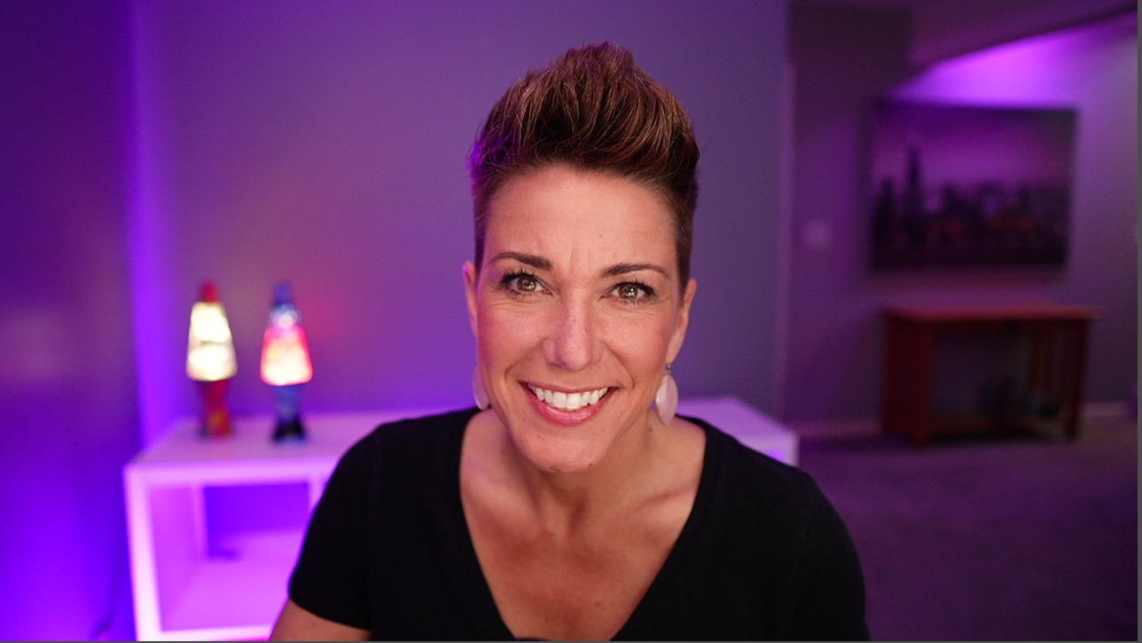 Portrait of Melanie Dyann Howe smiling against a purple background. There are lava lamps and a picture on the wall in the background.