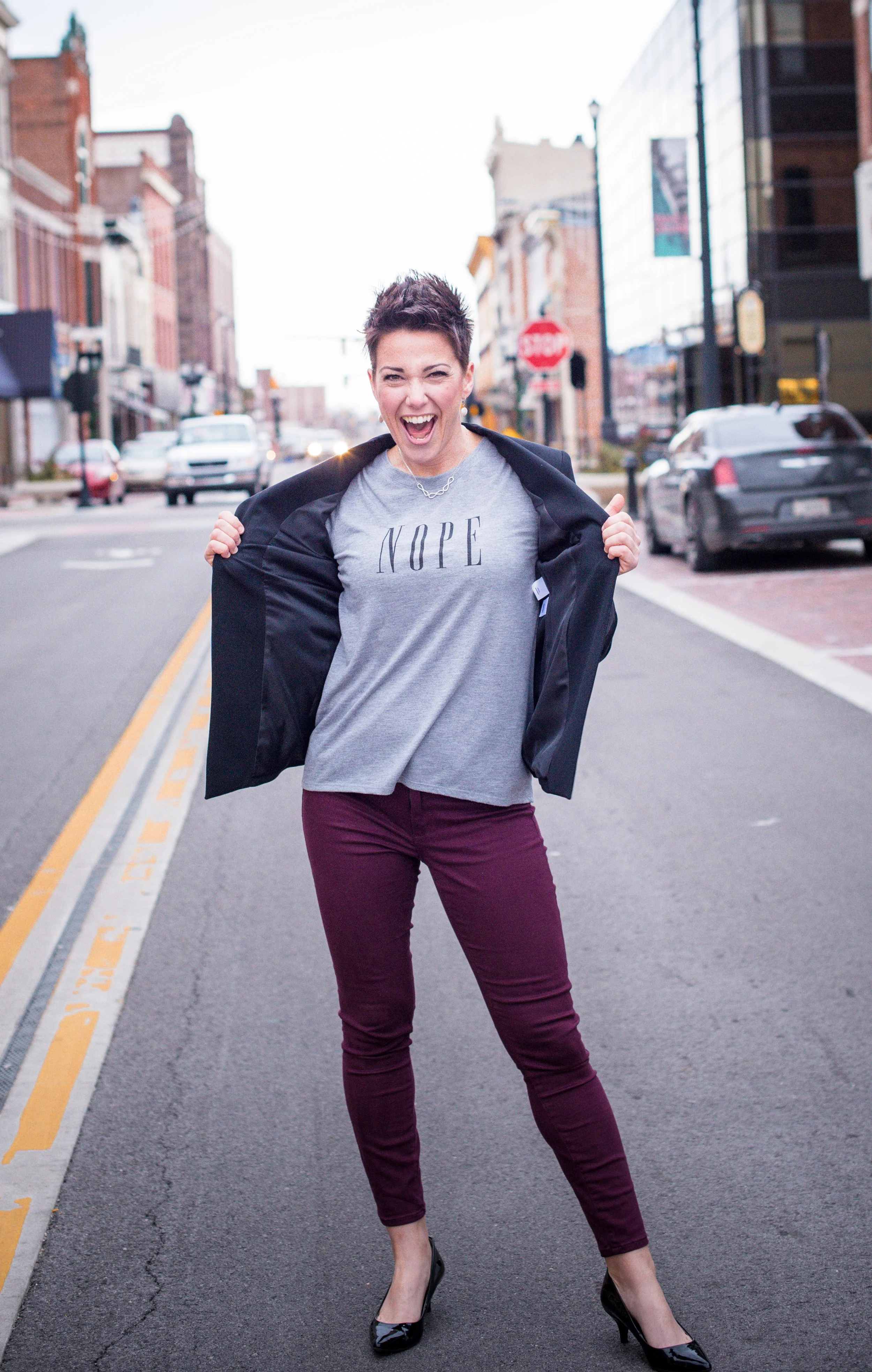 Mel standing on a city street, wearing a gray t-shirt with the word 'NOPE' printed on it. She is holding the blazer open with both hands and making a playful, enthusiastic expression.