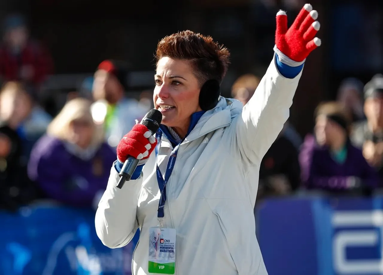 Mel announcing at the Indianapolis Monumental Marathon with a crowd in the background. She is wearing a VIP badge around her neck.