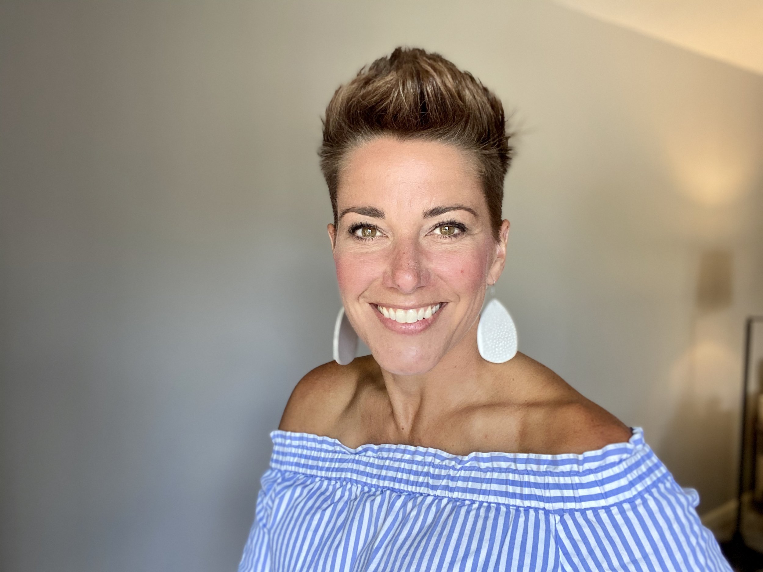 Melanie Dyan Howe wearing large white circular earrings and a blue and white striped off-the-shoulder top, standing in front of a plain wall.