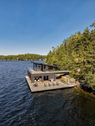 A modern house on a floating platform with a deck, surrounded by water and trees under a clear blue sky.