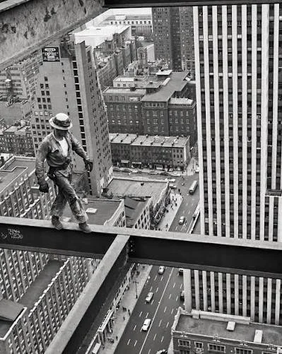 A construction worker walking on a steel beam high above a city street, surrounded by tall skyscrapers in black and white.