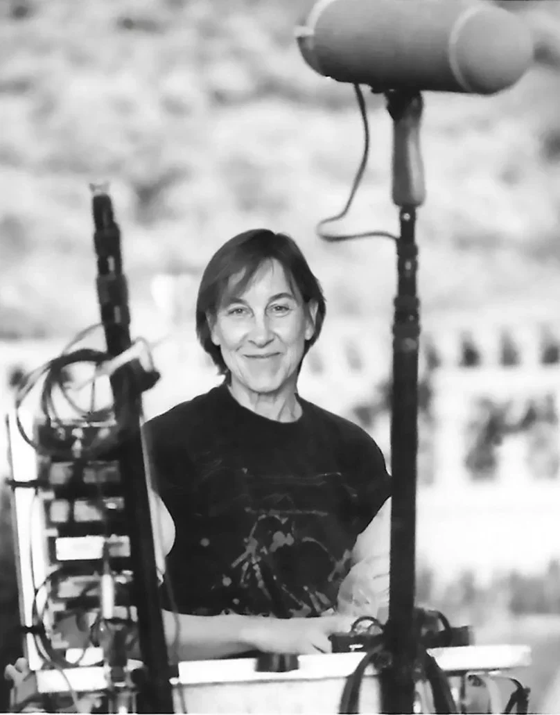 Patrushkha with short dark hair smiling at a camera, sitting outdoors surrounded by sound equipment on set.