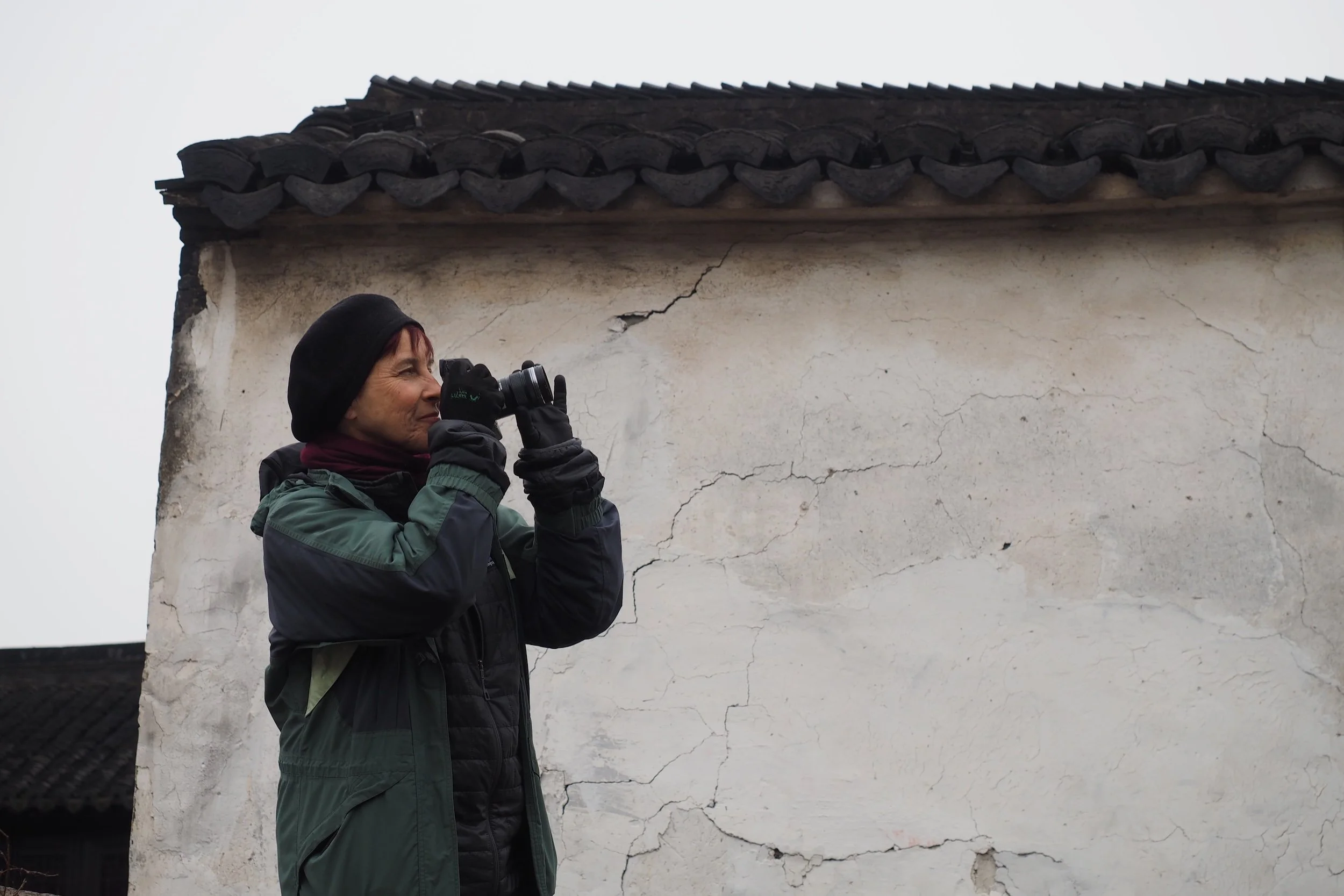 Patrushkha in outdoor clothing, wearing a black beanie and gloves, taking a photo with a camera against a weathered white wall with cracks, in an overcast setting.
