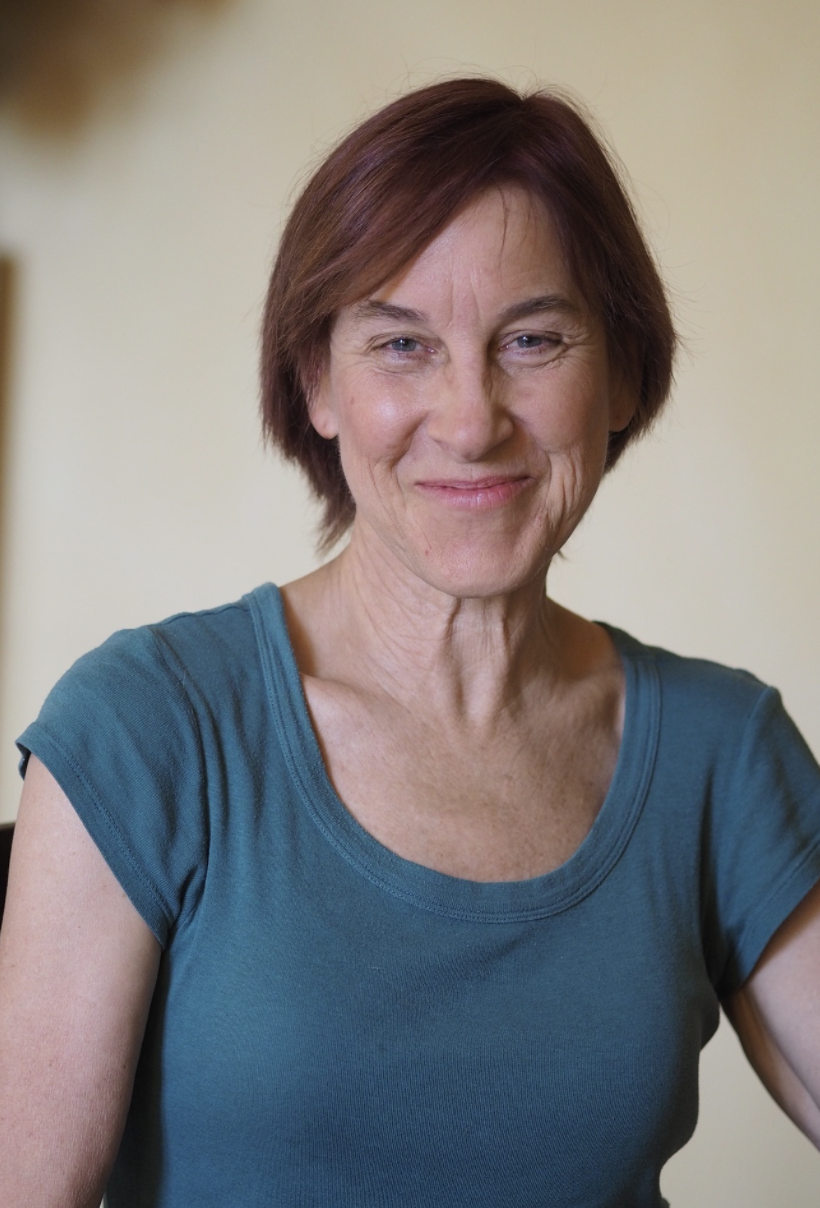 Patrushkha woman with brown hair and blue eyes, smiling slightly, wearing a teal short-sleeved shirt, sitting indoors against a neutral background.