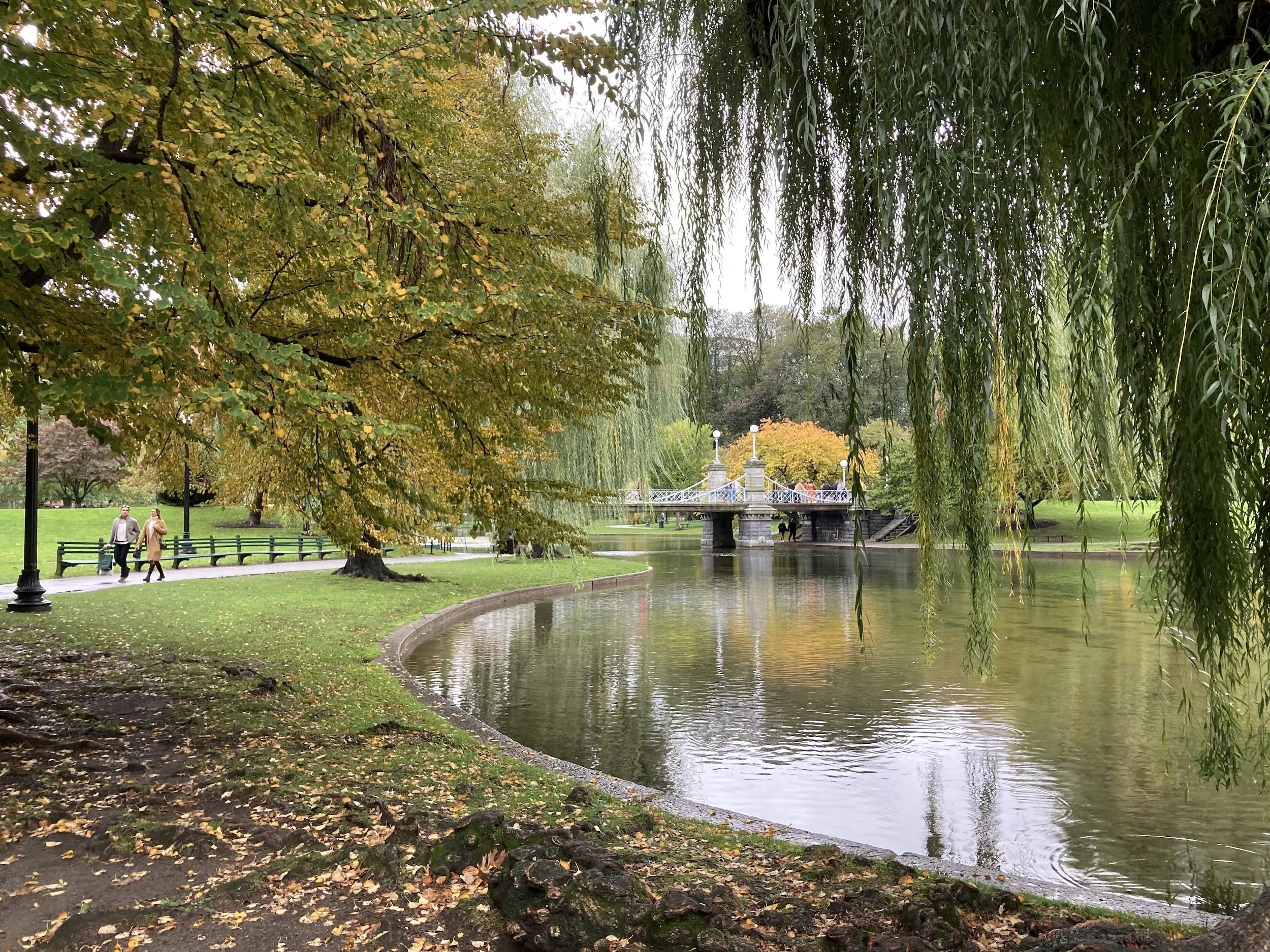 A park with trees near a pond, a bridge in the background, and two people walking on the path. The leaves are changing colors, indicating fall, with some orange and yellow leaves on the ground.