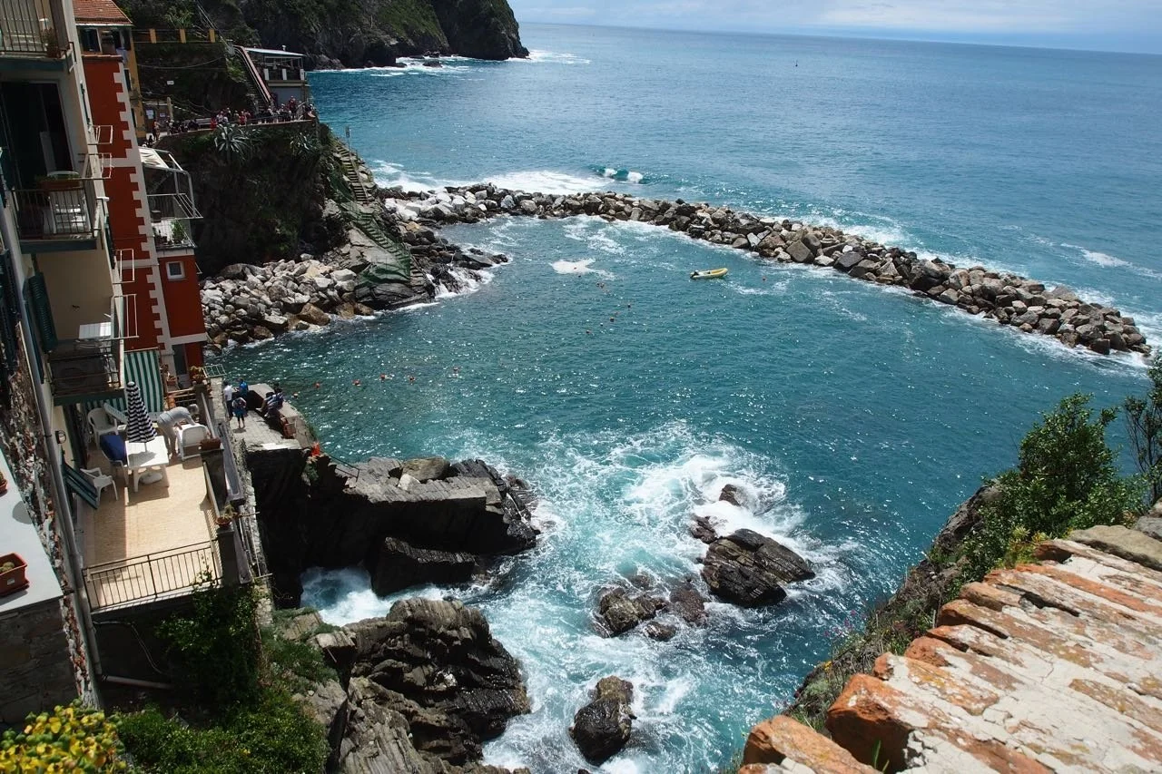 A coastline with buildings on the left and a rocky jetty extending into the ocean on the right, with the waves crashing against the rocks.