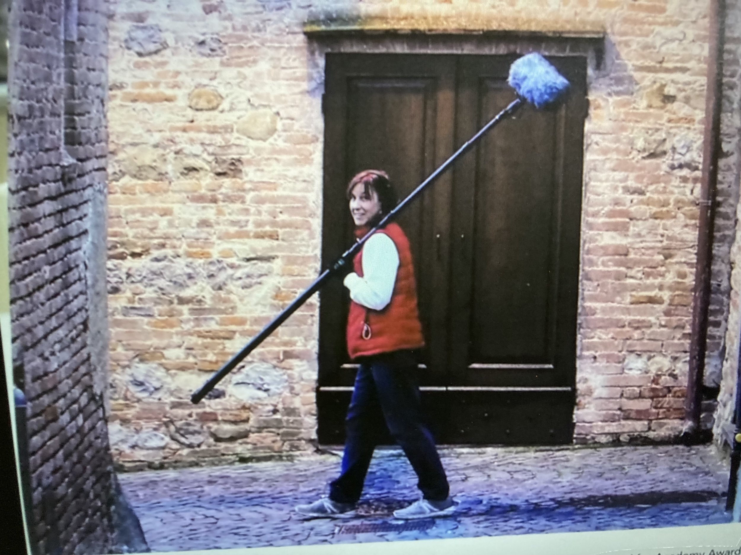 Patrushkha walking on a cobblestone street with a boom pole over her shoulder, standing in front of a brick wall with a black door.