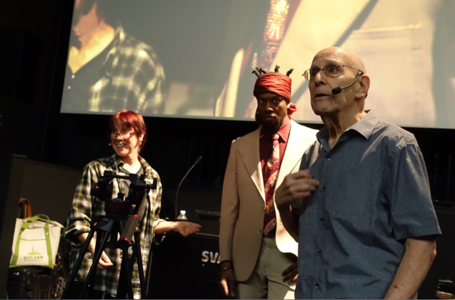 Patrushkha presenting on stage  smiling, another man in a beige suit with a red headscarf is looking serious, and an elderly man with glasses and a microphone headset is speaking or presenting.