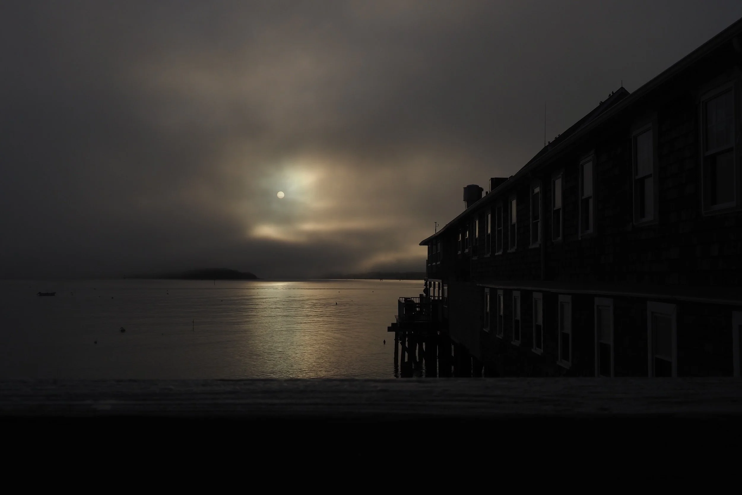 Dark, cloudy sky over a body of water, with the moon partially obscured, creating a reflective glow on the water's surface. On the right, there is a large pier on stilts extending into the water.