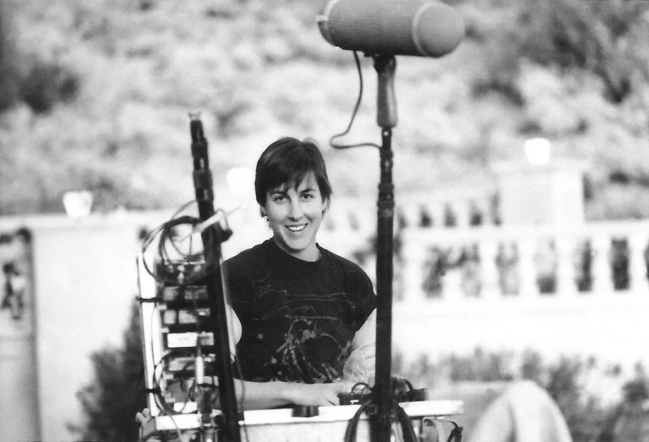 Patrushkha with short dark hair smiling at a camera, sitting outdoors surrounded by sound equipment on set.