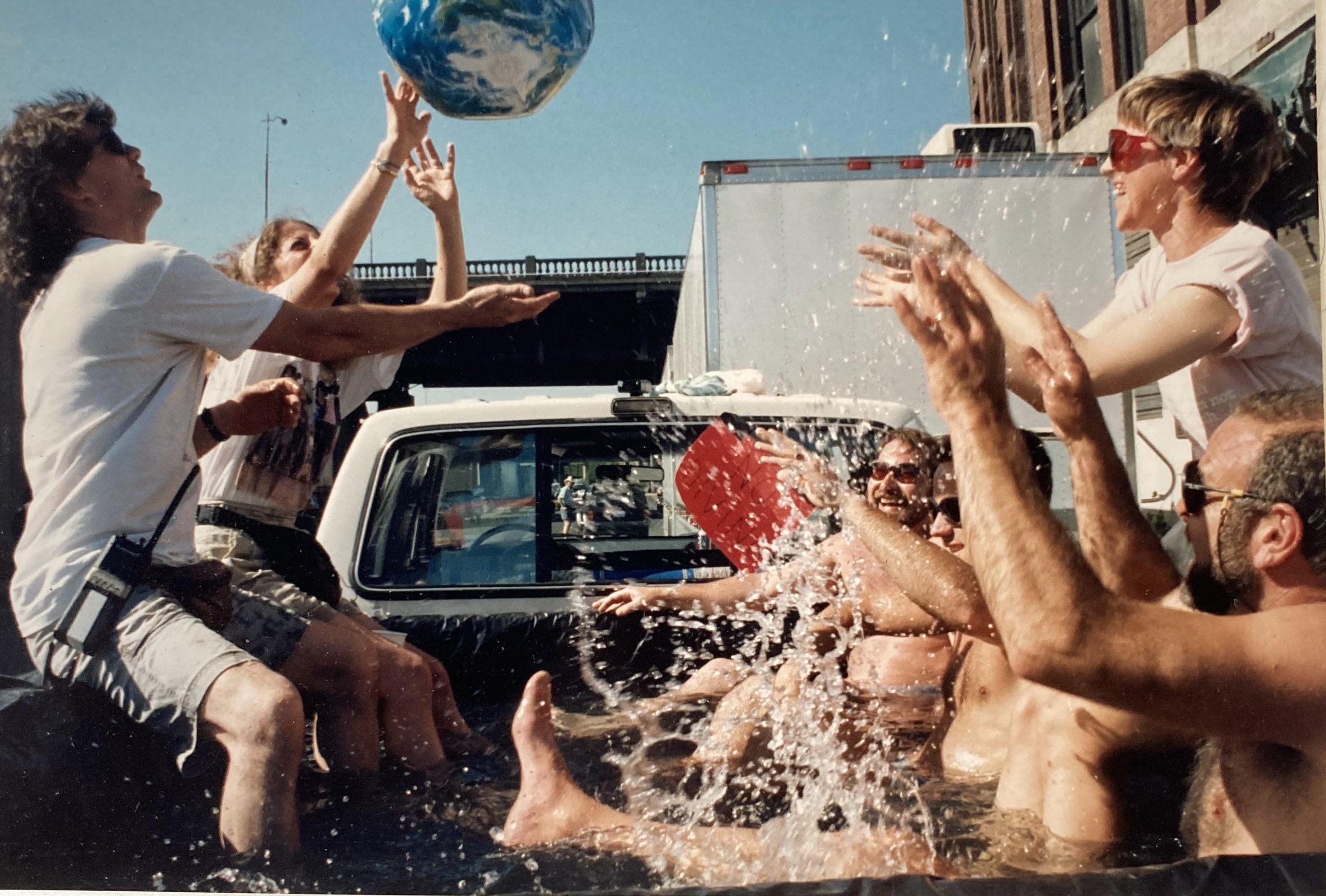 A group of people are gathered outside, throwing water and splashing each other during a playful activity on a sunny day.