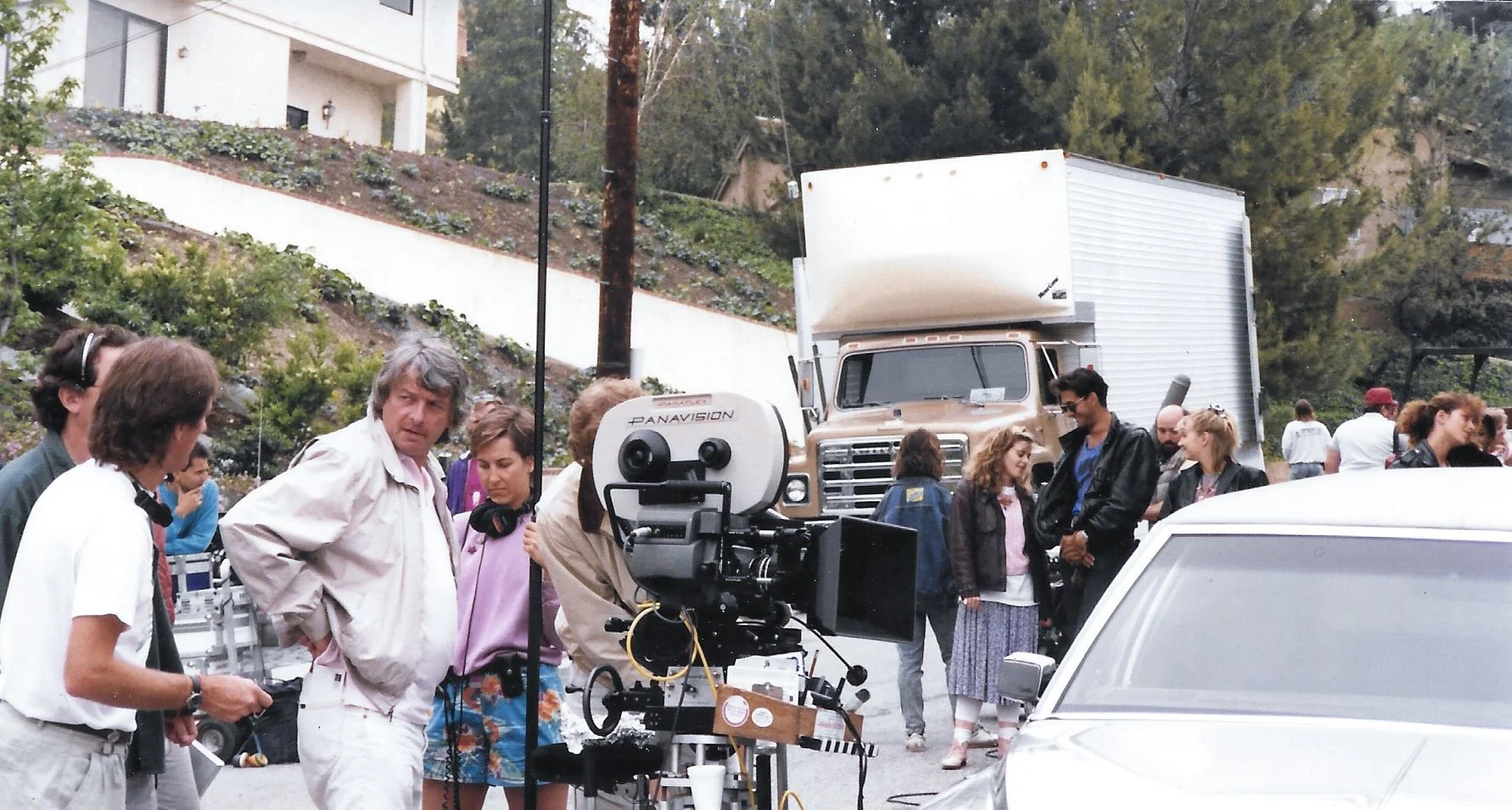 A group of people gathered outdoors around a film camera and production equipment. In the background, there is a large truck and a hillside with trees and buildings.