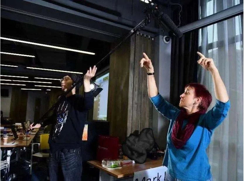 Patrushkha teaching students,  with one holding a boom microphone between them, both with arms raised and facing each other.