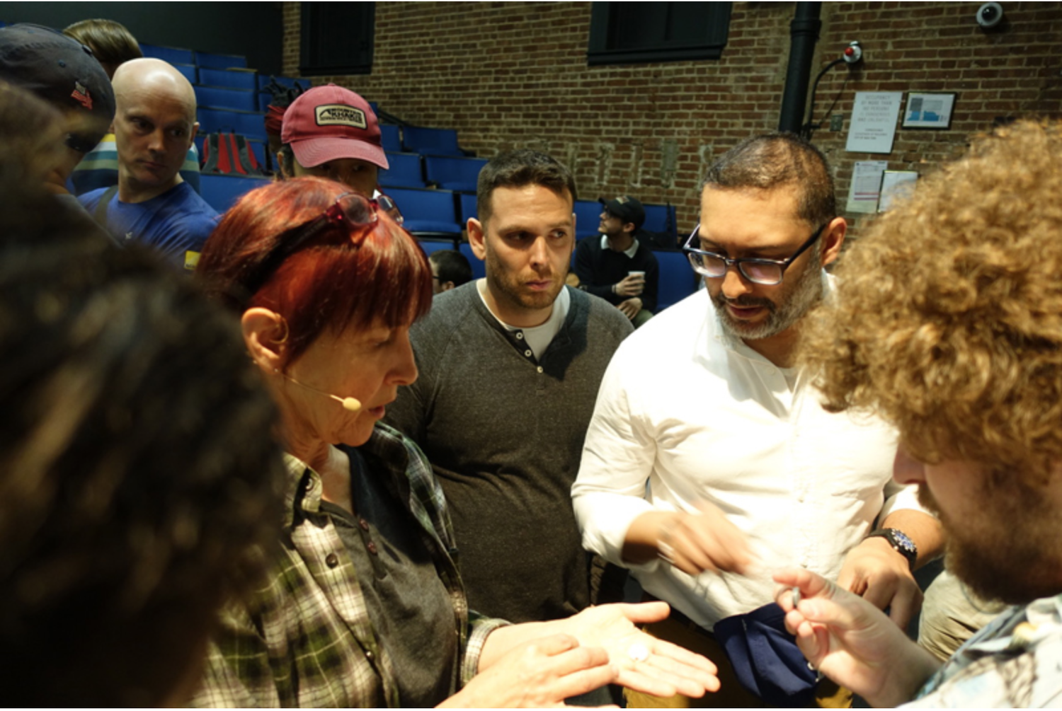 Group of people gathered around Patrushkha, showing and discussing an object, in a brick-walled indoor setting.