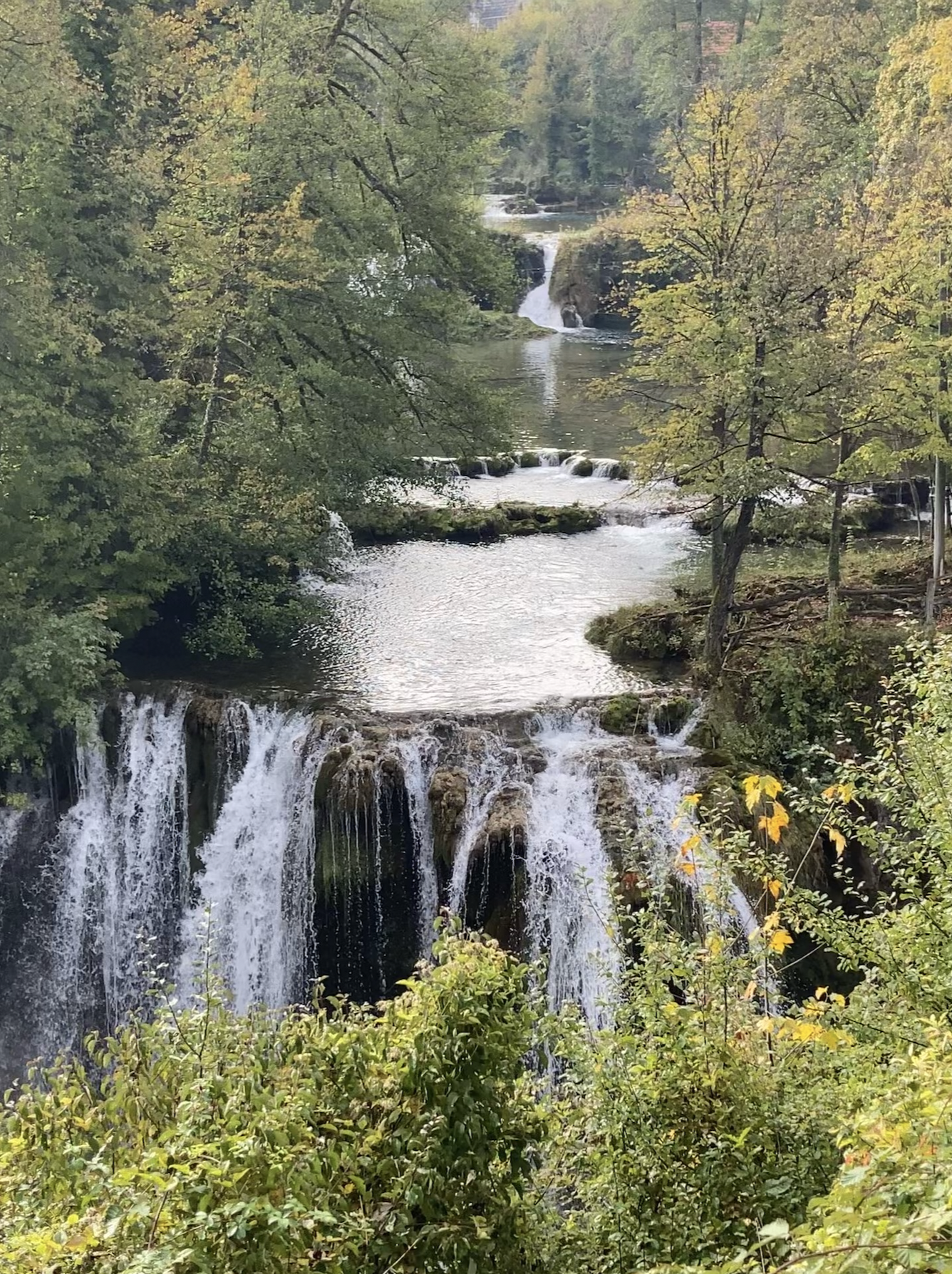 A scenic view of a multi-tiered waterfall surrounded by lush green trees, with a river flowing through a forested area.