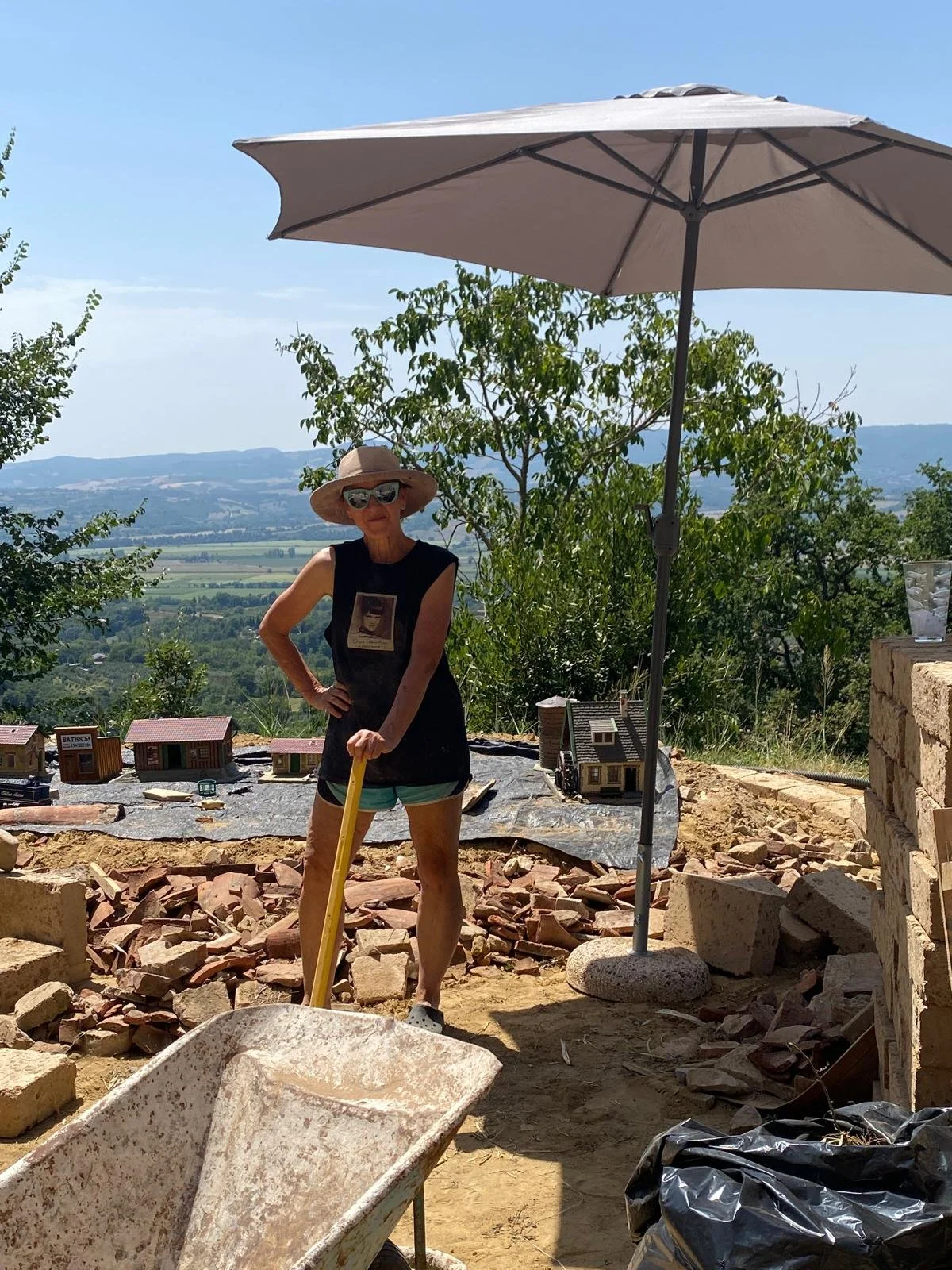 Patrushkha standing outdoors under a large patio umbrella during daytime, holding a shovel with a construction site surrounding her, small model houses on the ground behind her, and a scenic landscape with hills and trees in the background.