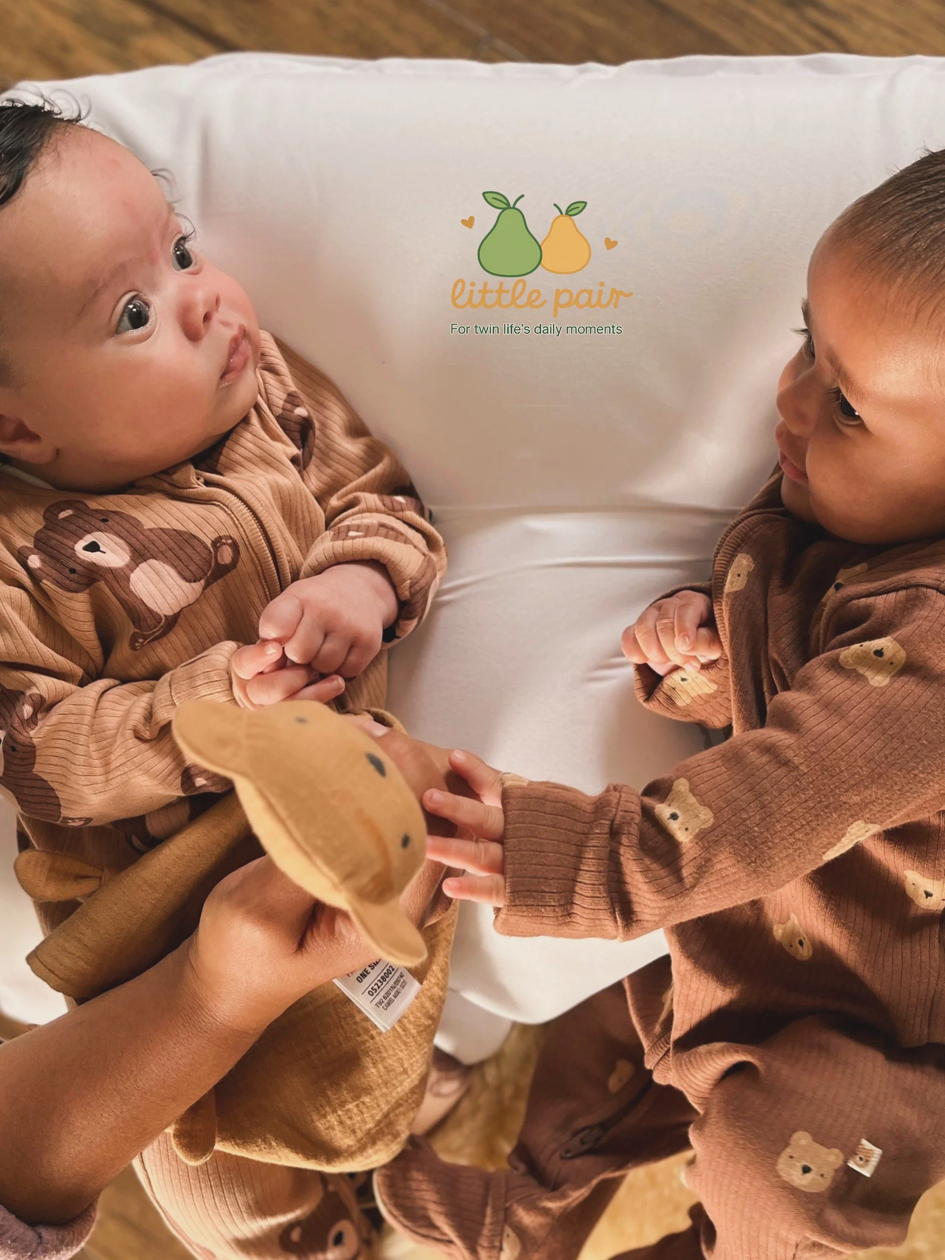 Twin babies lying side by side on a white support pillow during supervised playtime.